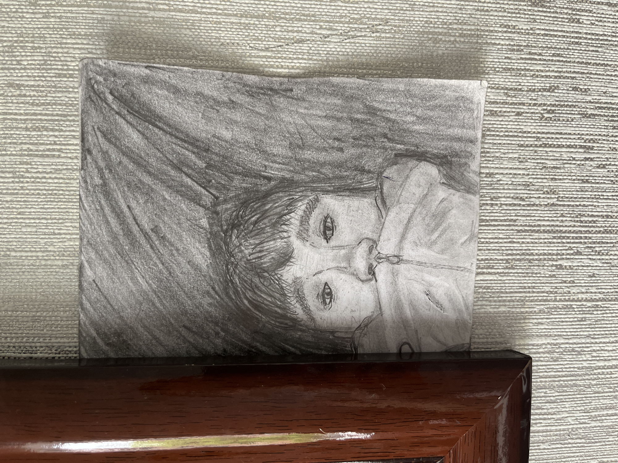 The image displays a framed pencil sketch of a person's face, partially obscured by their clothing. The sketch is positioned on a textured, light-colored wall. To the left of the sketch, a portion of a dark brown, glossy wooden frame is visible. The sketch itself is rendered in shades of gray, with visible pencil strokes creating texture and depth. The subject's eyes and nose are the most prominent features, with the rest of their face and body concealed. The background of the sketch is dark and abstract, with strong vertical lines. The overall lighting in the image appears to be natural, casting subtle shadows. There is no information in the image that would indicate its location in Saratov, Russia, or any specific city.