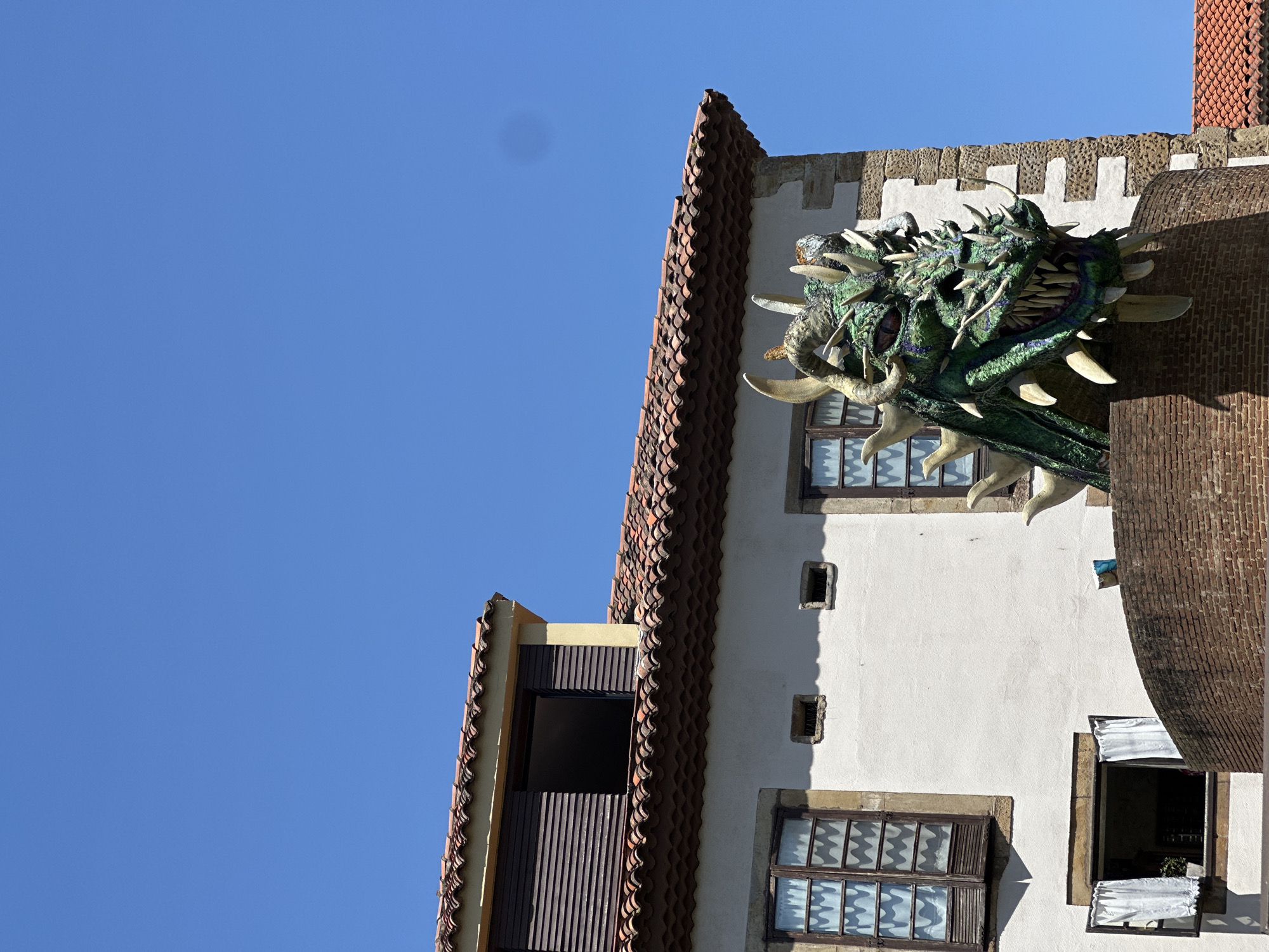 A low-angle shot captures the façade of a building in Gijón/Xixón, Spain, prominently featuring a dragon's head emerging from the wall. The building has white walls, brown-framed windows with wavy curtains, and a roof covered in orange-brown tiles. The dragon, which has horns and spikes on its head, appears to be bursting out from a brick pillar that extends vertically on the right side of the building. The sky is clear and blue.