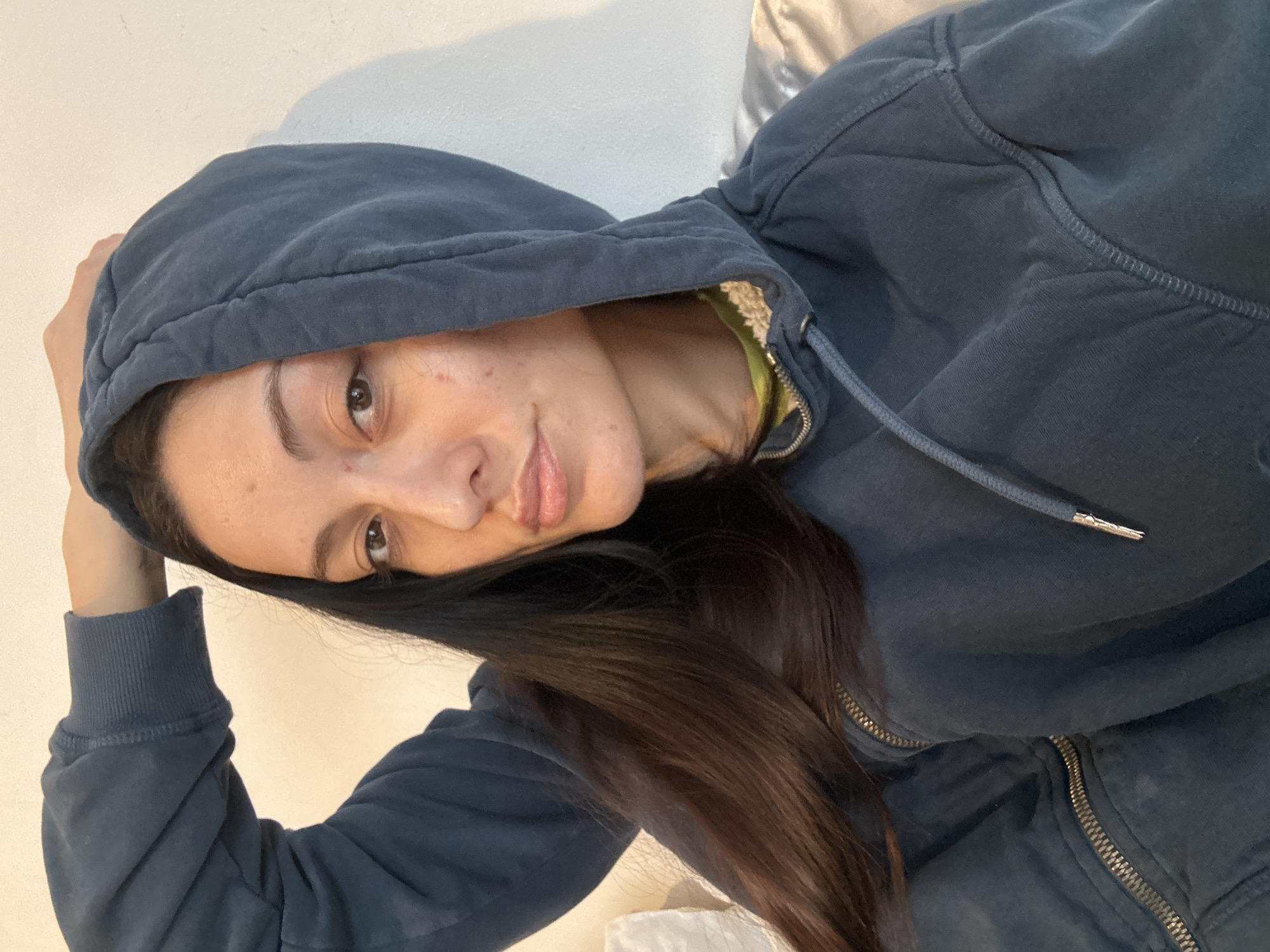 A close-up, tilted portrait captures a young person, likely female, wearing a dark blue or charcoal grey hooded sweatshirt. Her head is tilted to the left (viewer's right), and her gaze is directed towards the camera with a subtle, soft expression. She has long, dark brown hair that flows over her shoulder and chest, and light brown eyes. Her lips are naturally colored and slightly parted, and her skin shows natural textures and minor blemishes, indicating an unretouched appearance.
The person is dressed in a hooded sweatshirt that appears to be lined with a light-colored, possibly fleecy material inside the hood. A vibrant green or yellow t-shirt is visible underneath the zipped-up hoodie. The sweatshirt features a metal zipper down the front and a drawstring with silver aglets in the hood. Her right arm is raised, with her hand resting on the back of her head, contributing to the casual and relaxed pose.
The background is a simple, light-colored surface, most likely a white or light grey wall, suggesting an indoor setting. A shadow is cast on the wall behind her head, indicating a light source from the side. The overall impression is a candid and personal selfie. No text is visible in the image.
