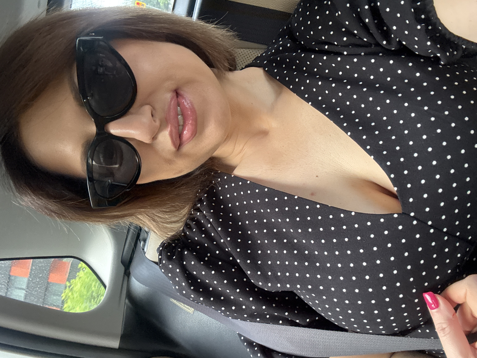 An image, rotated, depicts a woman from the chest up, positioned inside what appears to be a car, likely in Sofia, Bulgaria. She is wearing large black sunglasses and a black top or dress adorned with white polka dots. Her brown hair is visible around her face. Her lips are parted slightly, showing teeth, and are covered in a pink or mauve gloss. Her left hand, with a vibrant pink-red painted fingernail, is visible on the lower right, gently touching the fabric of her dress near her chest. A grey seatbelt crosses her lower torso. Through the car window in the upper left, a blurry view of outdoor scenery is visible, including green foliage and a building with reddish-brown panels and dark grey sections, suggesting an urban environment during daylight hours. The overall impression is a casual, relaxed pose. There is no visible text in the image.