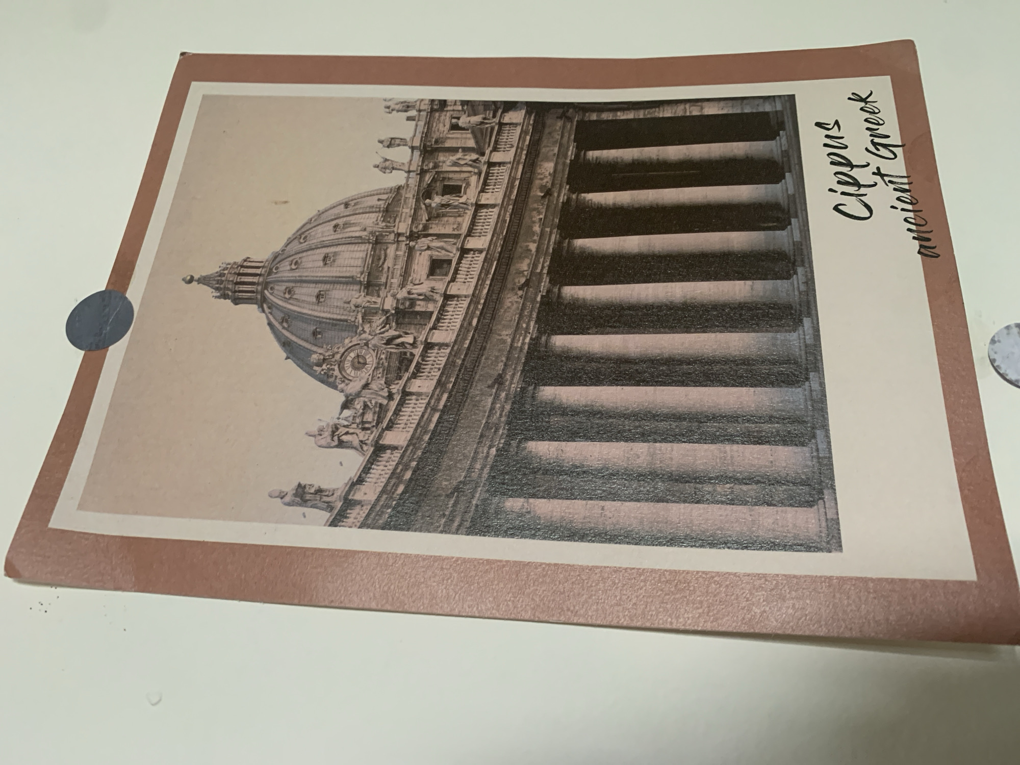 The image is a photograph of a postcard featuring a sepia-toned image of a grand classical building with a large dome and colonnade. The building appears to be St. Peter's Basilica in Vatican City, Rome, Italy, characterized by its distinctive dome and semicircular colonnade in the foreground. The postcard has a pink border and the words "Cippus ancient Greek" are handwritten in black script at the bottom right. A circular dark blue sticker is attached to the top right corner of the postcard. The postcard is leaning against a plain white wall. The location is not Kuala Pembuang, Indonesia.