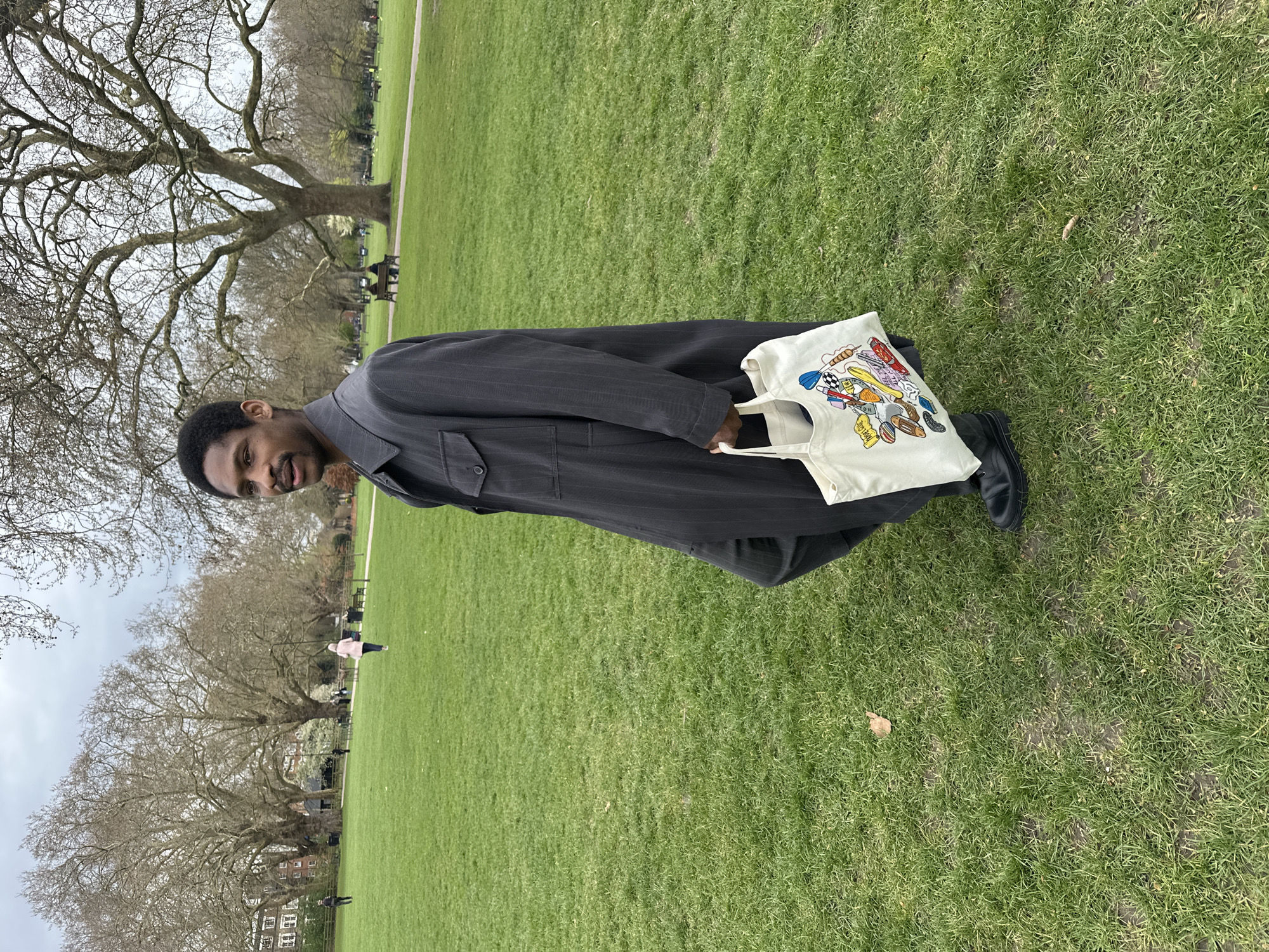 A man is standing in a grassy park, possibly in London, United Kingdom. He is wearing a long, dark-colored coat or overcoat and dark shoes. He is holding a white tote bag with colorful graphics. The park has large, bare trees, and other people can be seen in the background. The sky appears overcast.