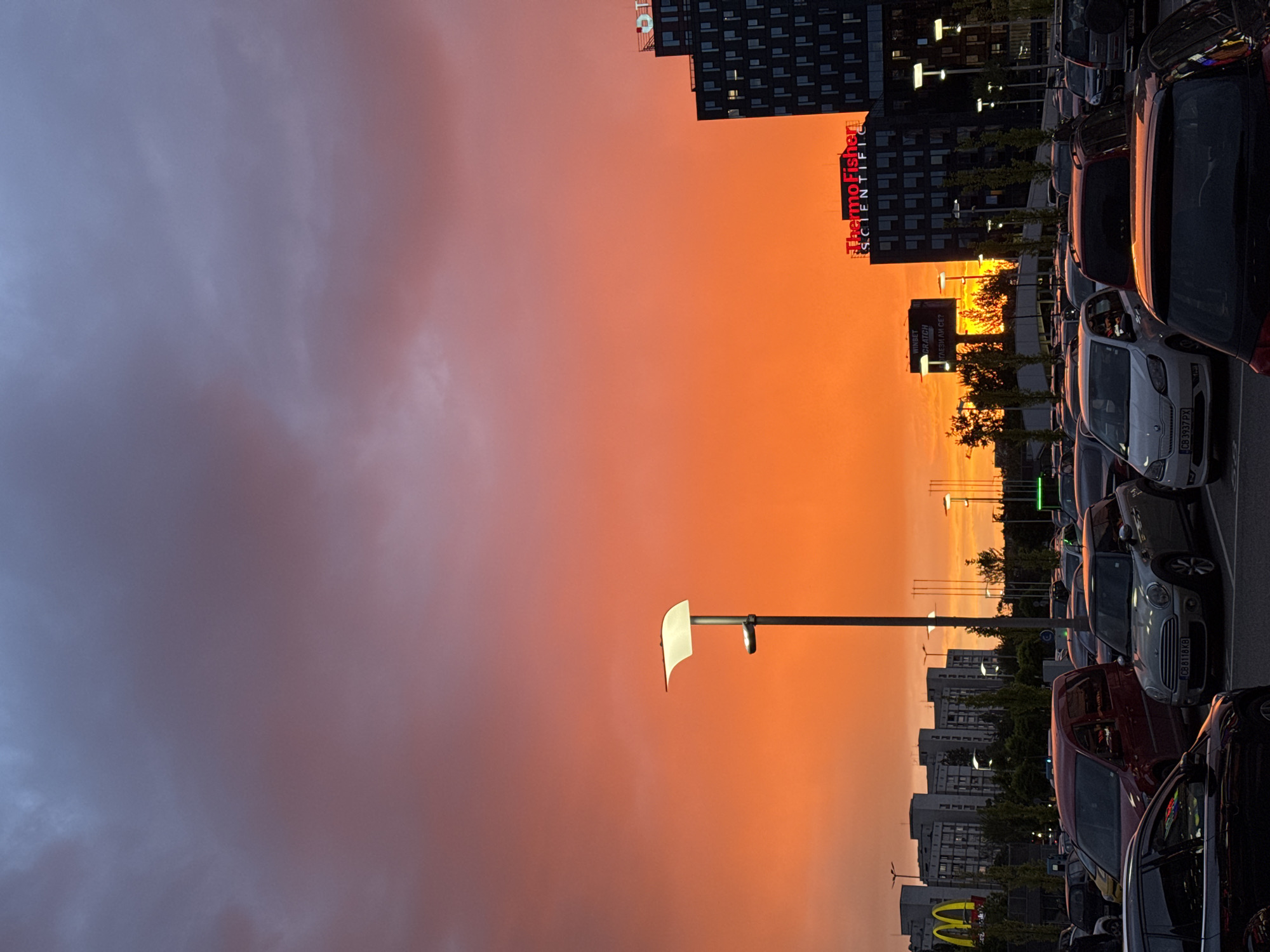 The image shows a parking lot in Sofia, Bulgaria, at sunset. The sky is a gradient of orange and pink. In the foreground are rows of parked cars. In the background, there are buildings, including one with a Thermo Fisher Scientific sign. There is also a McDonald's sign. The cars are of various makes and models. There are lampposts in the parking lot. The image is taken from a low angle.