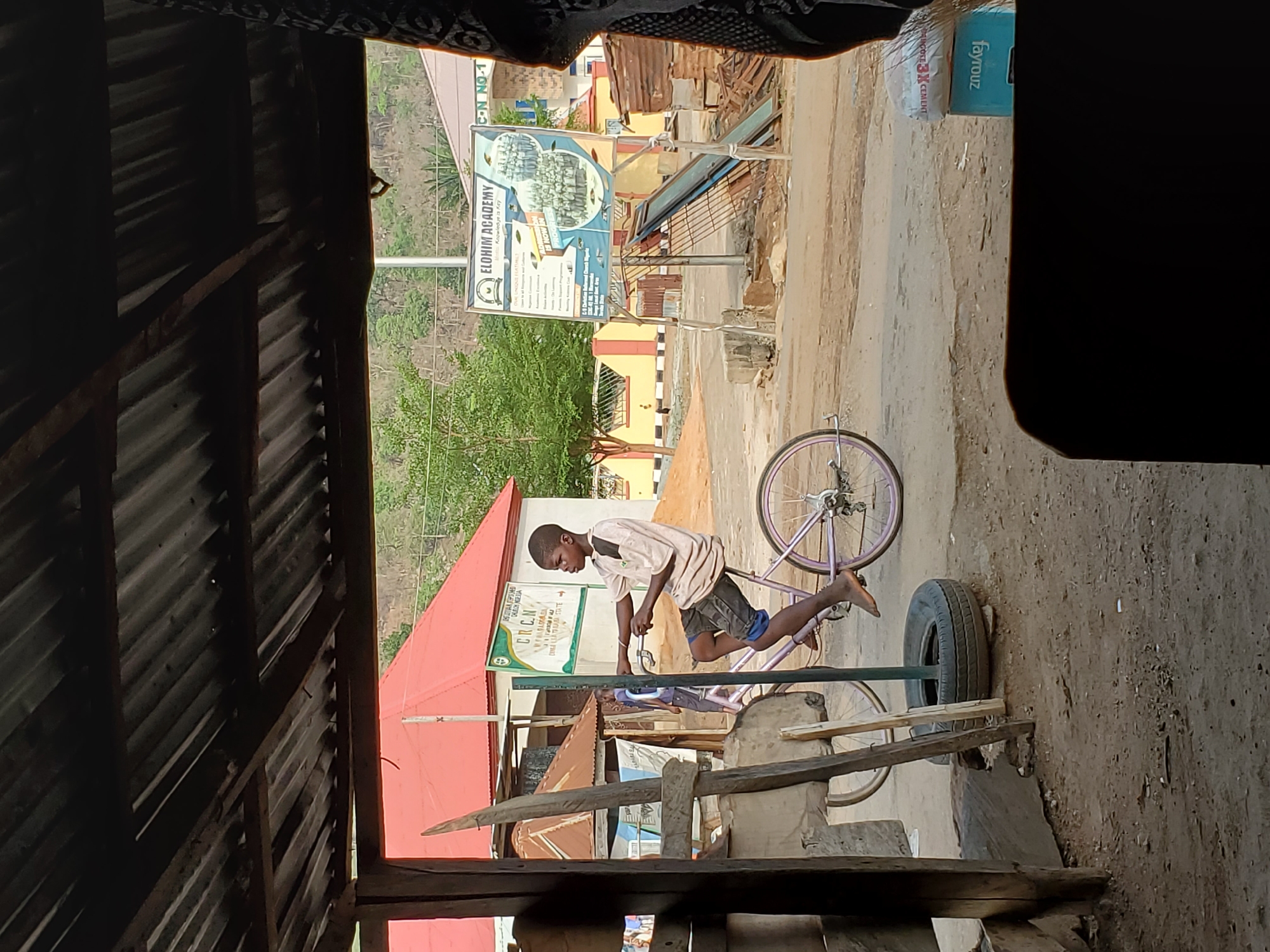A young boy in Mararaba, Nigeria, balances on a purple bicycle, standing on the frame instead of the pedals. He is in a residential area near Elohim Academy. There is a tire leaning against a wooden frame in front of him. In the background are buildings, a hill, and an Elohim Academy sign. The image is taken from inside a dark structure with corrugated metal sheets.