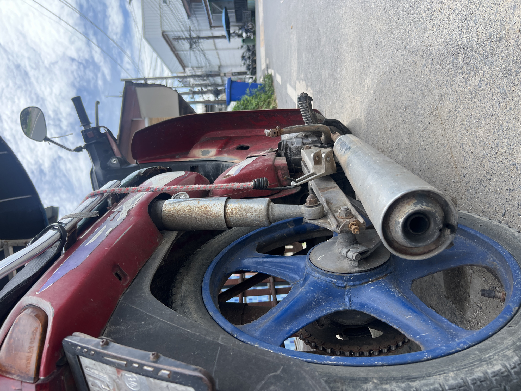 This is a close-up, low-angle shot of the rear wheel and exhaust of a red motorcycle parked on an asphalt street. The motorcycle's blue rim is prominent, with a visible chain and sprocket. The exhaust pipe is silver and appears slightly rusted. In the background, buildings with power lines and a blue trash bin are visible, suggesting an urban or semi-urban setting. A person is not clearly visible, but a motorcycle with a rider is in the far background, suggesting a street in Choeng Thale, Thailand. The sky is cloudy.