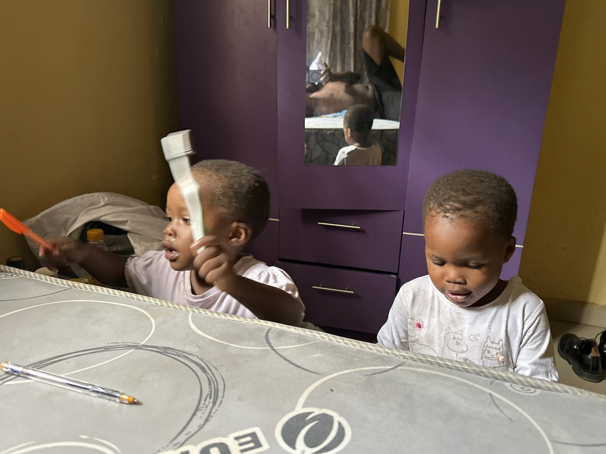 Two young children, a boy and a girl, are in a room in Narayi, Nigeria. The boy, on the right, wears a white t-shirt with cartoon animal graphics and is looking down. The girl, on the left, is facing right and holding a white object, possibly a toothbrush, and an orange object, possibly a toy. Both children are seated at a table with a grey patterned surface. In the background, a large purple wardrobe with gold handles is visible. A mirror on the wardrobe reflects a person lying down and another child standing. The room has yellow walls.