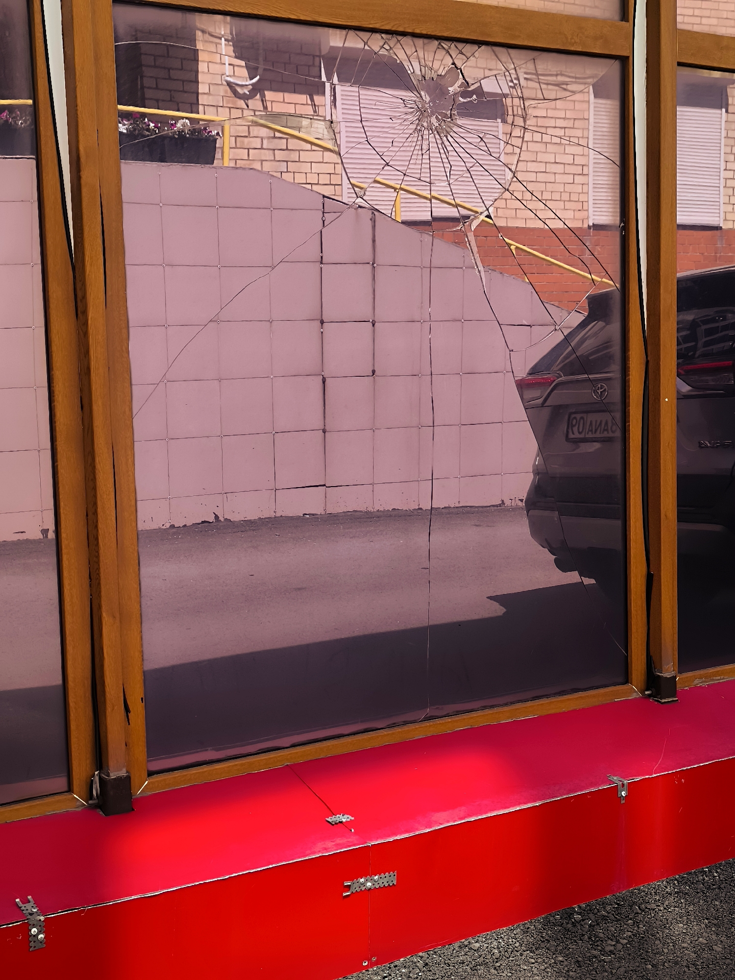 A cracked windowpane is visible in the foreground, reflecting a street scene. The window has a wooden frame and is set into a bright red structure. The reflection shows a brick building with barred windows, a yellow railing, and a car with a license plate that reads "PO AMAS". The overall impression is that of a damaged storefront or window. The location of Karaganda, Kazakhstan, is not evident from the image itself and would require external information to confirm.