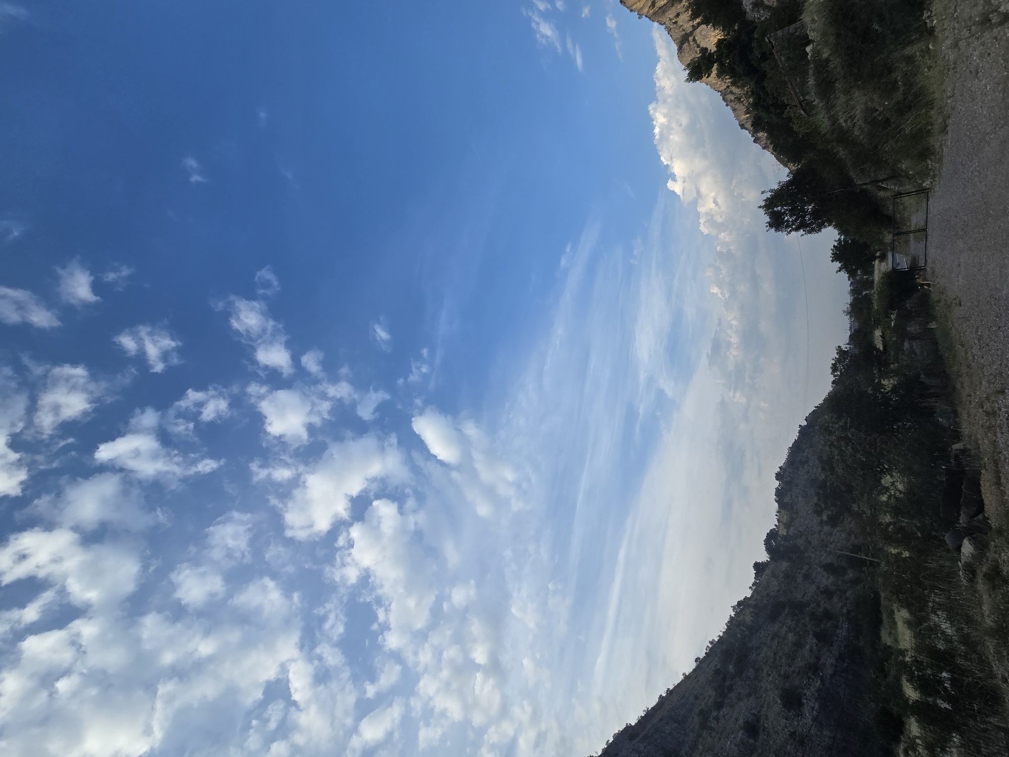 The image depicts a natural landscape with rugged terrain and a bright blue sky dotted with white clouds. On the left side, a steep, dark, rocky hillside covered in sparse vegetation slopes downwards. On the right, another rocky slope rises, also sparsely vegetated. In the foreground, a gravel path leads into the scene, marked by a metal gate. Beyond the gate, trees and low-lying vegetation are visible, suggesting a rural or semi-rural area. The lighting indicates it is daytime, possibly late afternoon or early morning due to the angle of the sun and the contrast in the sky. The environment suggests a mountainous or hilly region with limited human development. This appears to be a location in the outskirts of Podgorica, Montenegro, possibly in a natural reserve or a less developed area leading into the hills surrounding the city. There are no people or animals clearly visible in the main frame of the image, although a small, indistinct animal may be present near the gate.