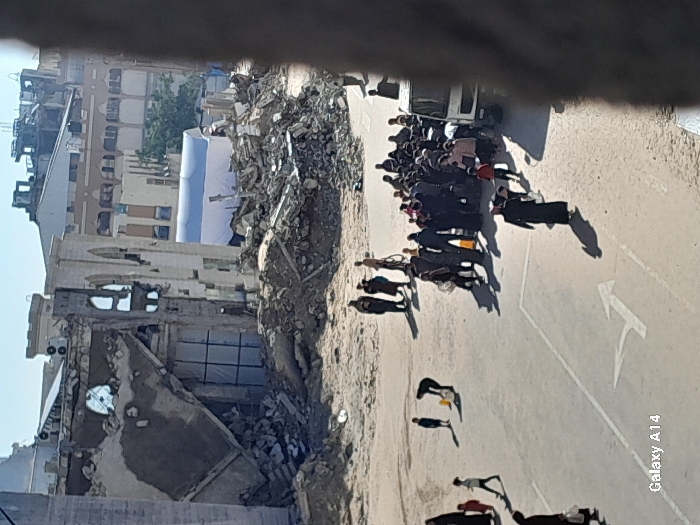 This image captures a wide view of a heavily damaged urban area in Khan Yunis, Palestinian Territory, under clear daylight.
The scene is dominated by widespread destruction, with a large, multi-story building on the left exhibiting extensive structural collapse; its upper section is partially destroyed, exposing internal structures and debris, while remnants of arched window frames and decorative elements are visible. To the right of this building and extending across the midground are vast piles of rubble and debris. In the background, beyond the rubble, a white, tent-like structure is visible, with additional damaged buildings further in the distance under a clear blue sky.
In the foreground and midground, a wide, open area, appearing to be an unpaved or dirt-covered former street or plaza, features faint white painted road markings, including a prominent arrow indicating a left turn and straight direction.
Multiple individuals are dispersed across this open area. On the far left, a woman in a black abaya walks with two children. Slightly to their right, a child stands near an adult bent over, appearing to interact with a yellow bucket or container on the ground. Further back, a small group of 3-4 adults is present, one with a bicycle. Towards the right midground, a dense cluster of approximately 30-40 individuals, comprising adults and children, is gathered. Many are dressed in dark attire, and they are positioned near the front of a white light truck or van, which is partially visible on the far right edge of the group. Another individual in a long, dark garment walks towards the bottom right, away from the main crowd.
Bright sunlight illuminates the scene, casting long, distinct shadows of the people and objects towards the lower left.