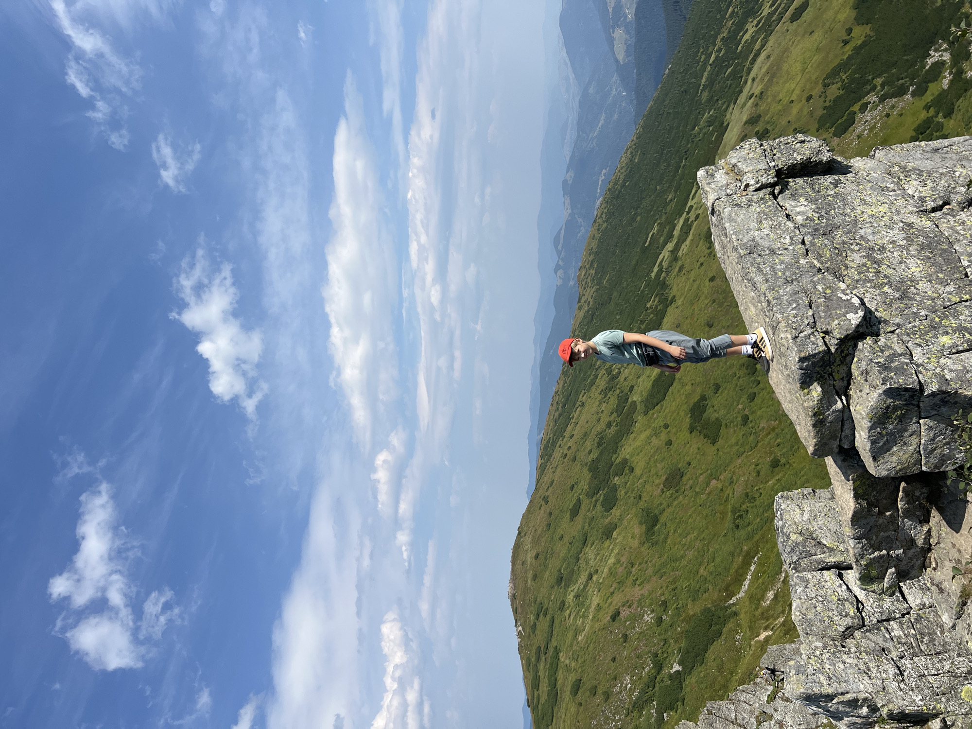 In the city of Vorokhta, Ukraine, a young boy wearing a red cap, a green t-shirt, and gray pants stands on a rocky outcrop, looking towards the camera. He is positioned on the right side of the image, with a vast mountain landscape stretching out behind him. The mountains are covered in lush green vegetation, with a winding path visible on one of the slopes. The sky above is a clear blue, dotted with scattered white clouds. The boy's posture suggests a moment of rest or admiration of the scenery.