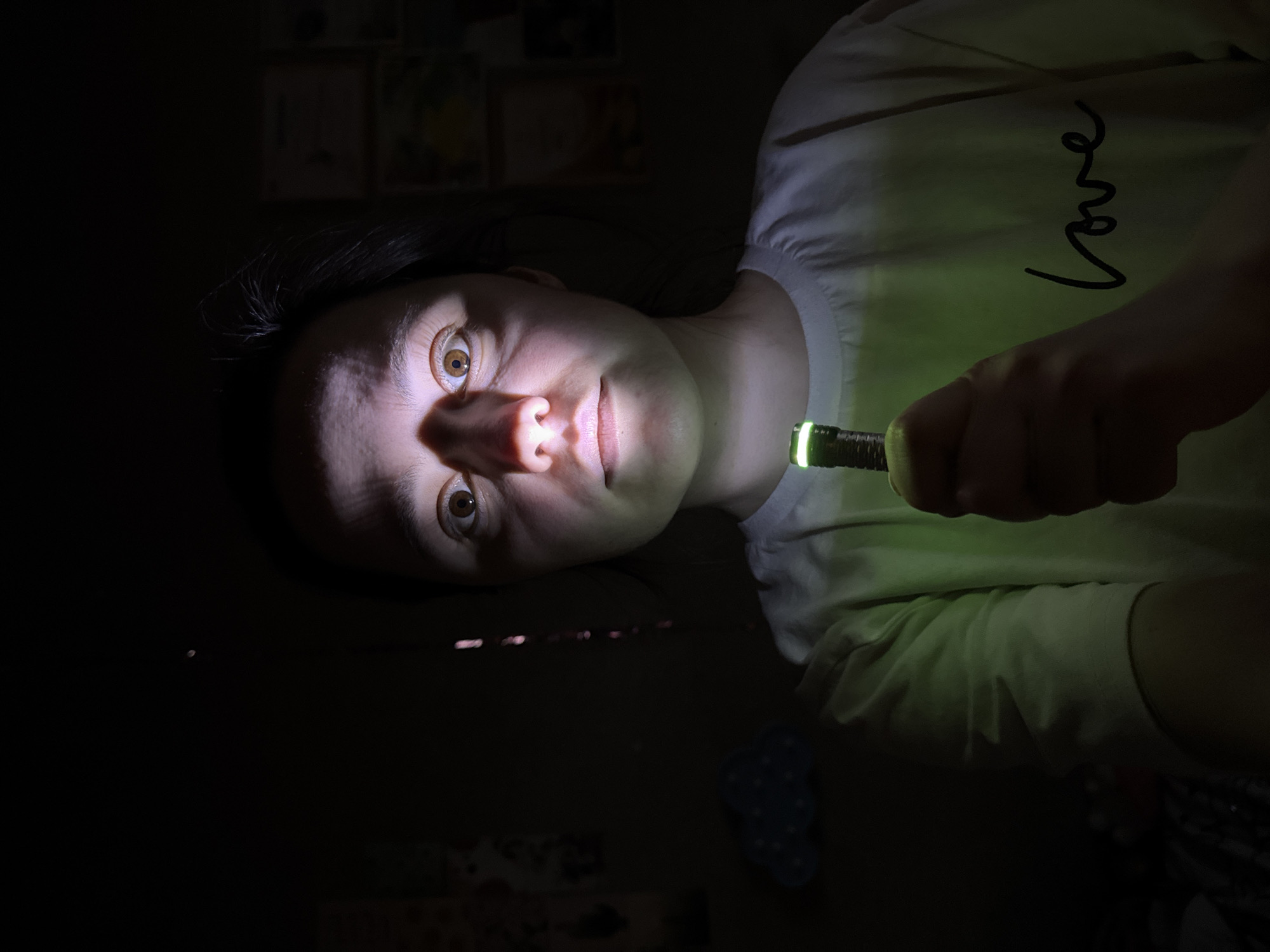 A low-angle, close-up shot depicts a person holding a flashlight that illuminates their face in a dark room. The person, who has light skin and brown eyes, is wearing a white t-shirt with the word "love" written on it. The flashlight casts a strong beam of light onto their face, creating harsh shadows. The background is dark and mostly indistinct, though some shapes and patterns are vaguely visible, including a partially visible blue cloud. The location of the image is unknown.