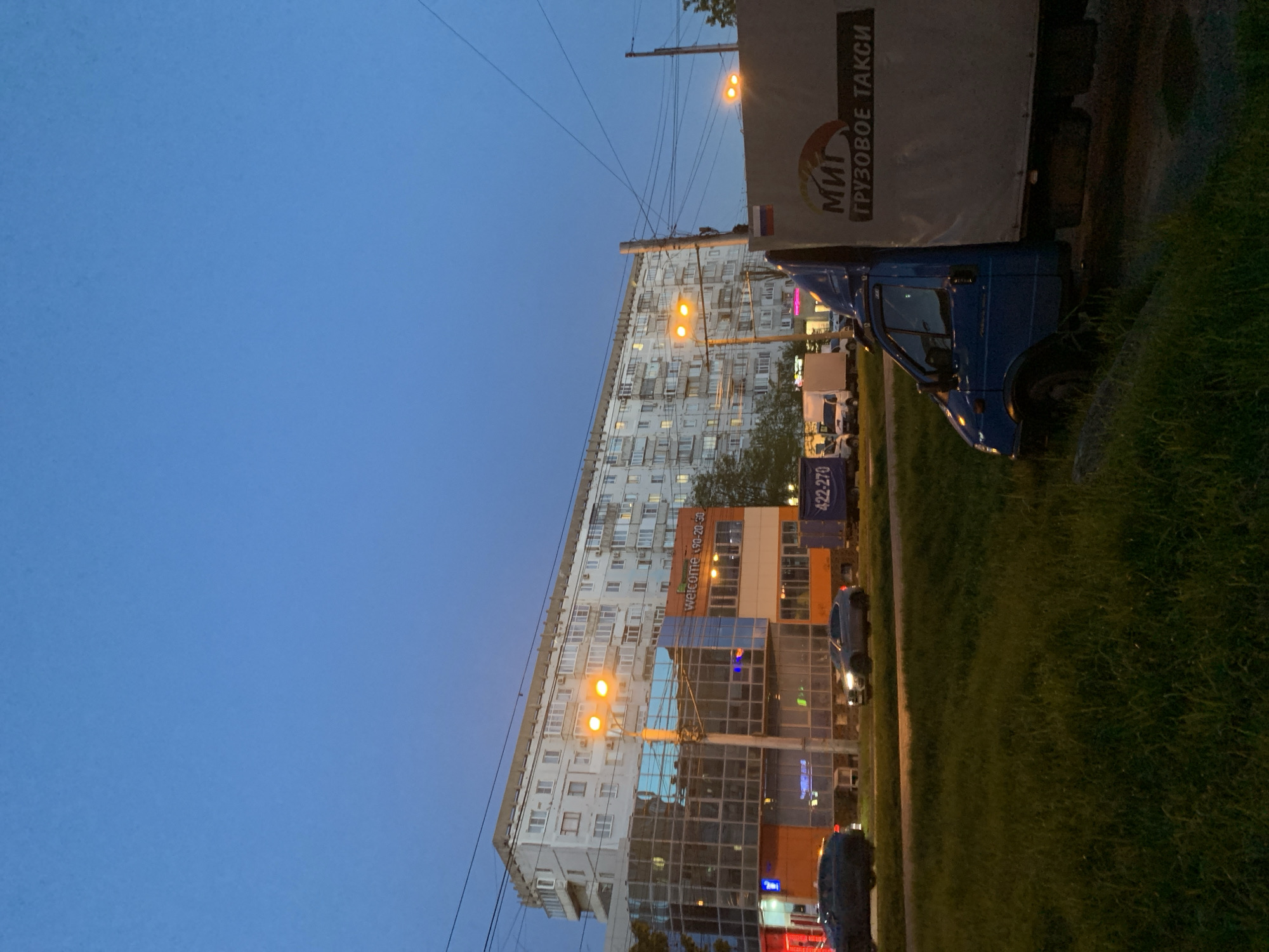 The image captures an urban street scene at dusk or early evening, characterized by a deep blue sky and artificial lights illuminating buildings and vehicles. The overall perspective is slightly rotated counter-clockwise.
In the background, a tall, multi-story residential apartment building dominates, its facade featuring numerous windows, some of which are lit from within. Below this, a modern commercial building with large glass windows and orange-colored panels is visible, reflecting the evening light. Several streetlights emit a warm, orange glow, contrasting with the cool blue of the sky and casting light on the surroundings. Utility wires crisscross the sky.
In the foreground to the right, a blue truck with a white cargo box is prominent, partially obscured by a grassy verge. The side of the cargo box features a logo and text in Cyrillic, reading "МИТ ГРУЗОВОЕ ТАКСИ" (MIT Cargo Taxi), with a small Russian flag emblem. Other vehicles, including cars and possibly other trucks, are visible further down the street and in the background. A large patch of green grass occupies the lower right portion of the frame.
Visible text on the commercial building includes "WELCOME" and phone numbers "190-20-50" and "422-270". The scene depicts typical city life in the evening, consistent with an urban setting like Stavropol, Russia, with vehicles either parked or in motion. No people are clearly discernible.
