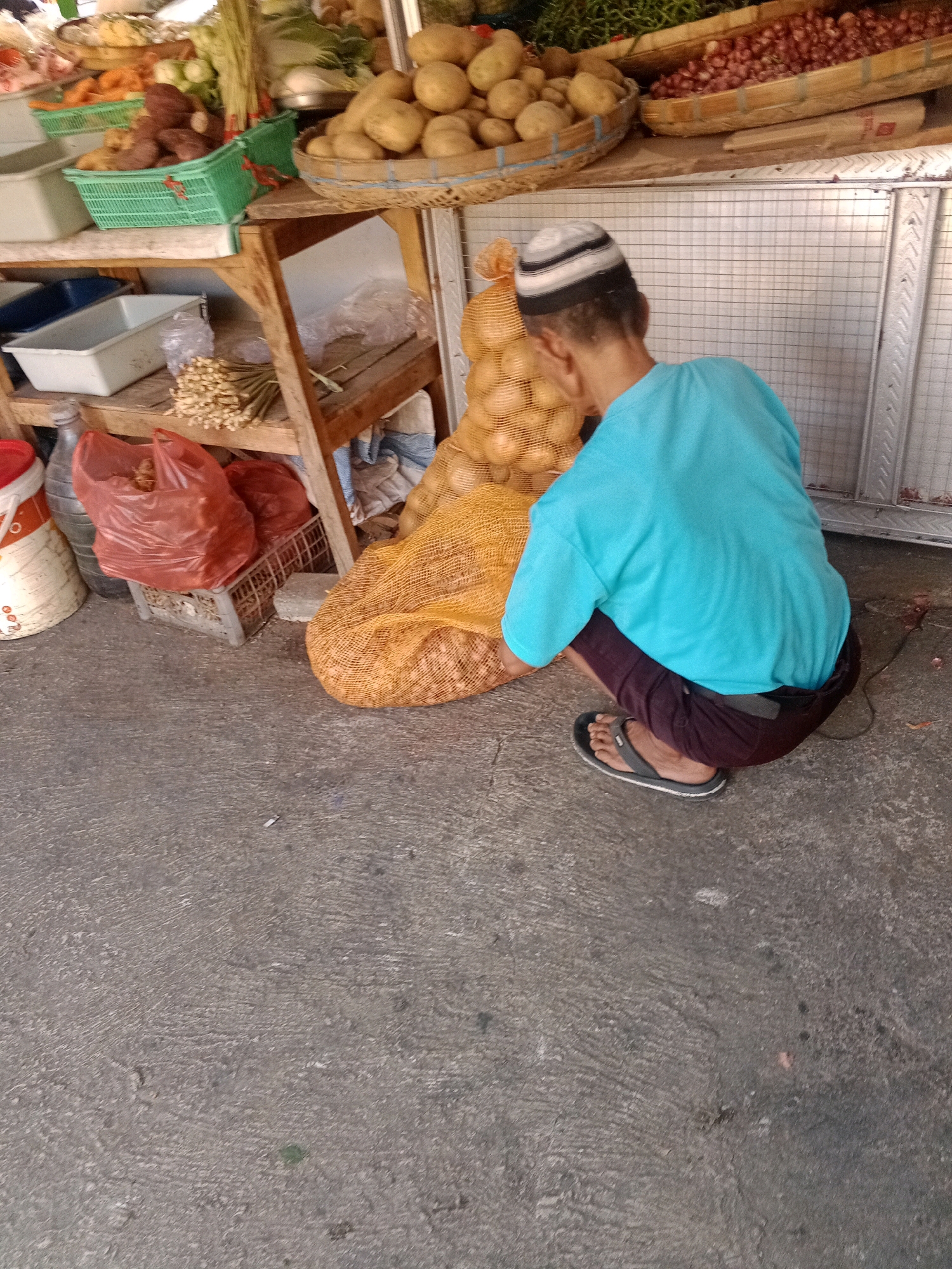 In Wonogiri, Indonesia, a person wearing a blue t-shirt and a striped cap is crouching next to a large, netted sack filled with potatoes. The sack is orange and sits on a concrete ground surface. Behind the person, shelves display various produce, including potatoes, onions, and what appear to be carrots and leafy greens, some contained within green baskets. Several plastic bags, one red and one clear, and a water bottle are also visible on the lower shelf, along with a white bucket with orange markings. The scene takes place outdoors, possibly at a market stall, with a metal grid wall in the background.