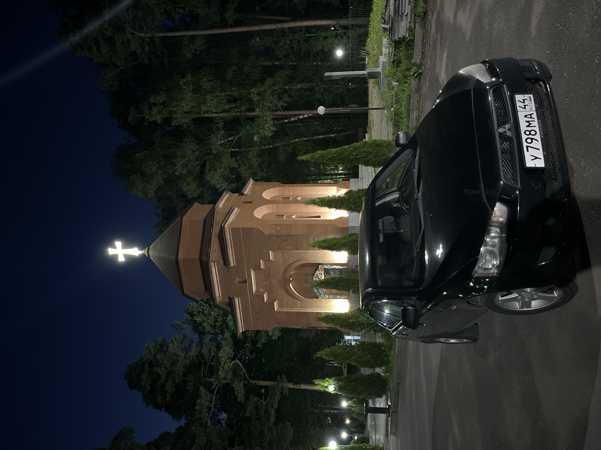 A black Mitsubishi Lancer is parked in front of a light brown, stone-clad building that appears to be a small chapel or monument, illuminated by lights. The building has arched doorways and a cross topped with a bright light is visible on its roof. The scene is set at night, with dark trees and a clear, dark blue sky in the background. Several small, conical evergreen trees are planted in a row leading up to the building. Streetlights are visible on the left side of the image, and a fence can be seen in the background on the right. The license plate of the car reads "Y 798 MA 44" and "РОССИЯ". The location is identified as the Klyazmensky Church of the Resurrection in Kostroma, Russia.