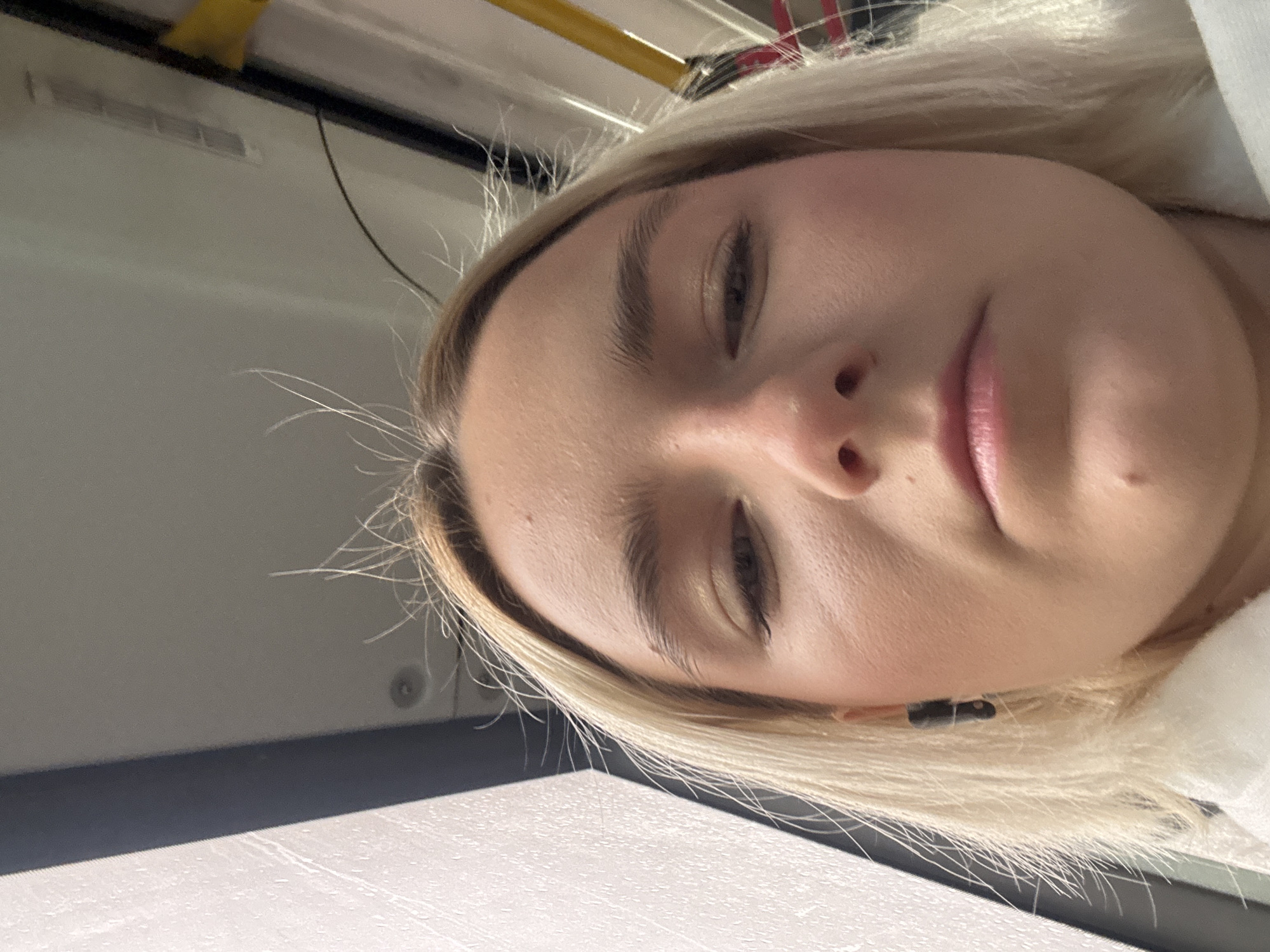 A close-up, angled self-portrait of a light-skinned young woman with blonde hair. She has fair skin, light eyebrows, and closed eyes. Her lips are a pale pink. Part of her neck and shoulder are visible, covered in white fabric. A small black object is visible in her blonde hair near her neck. The background is comprised of the interior of a vehicle (possibly a train), indicated by the presence of a white ceiling panel, a black frame, a yellow object attached to a bar, and a vent. No definitive features identifying Saint Petersburg, Russia are visible in this image.