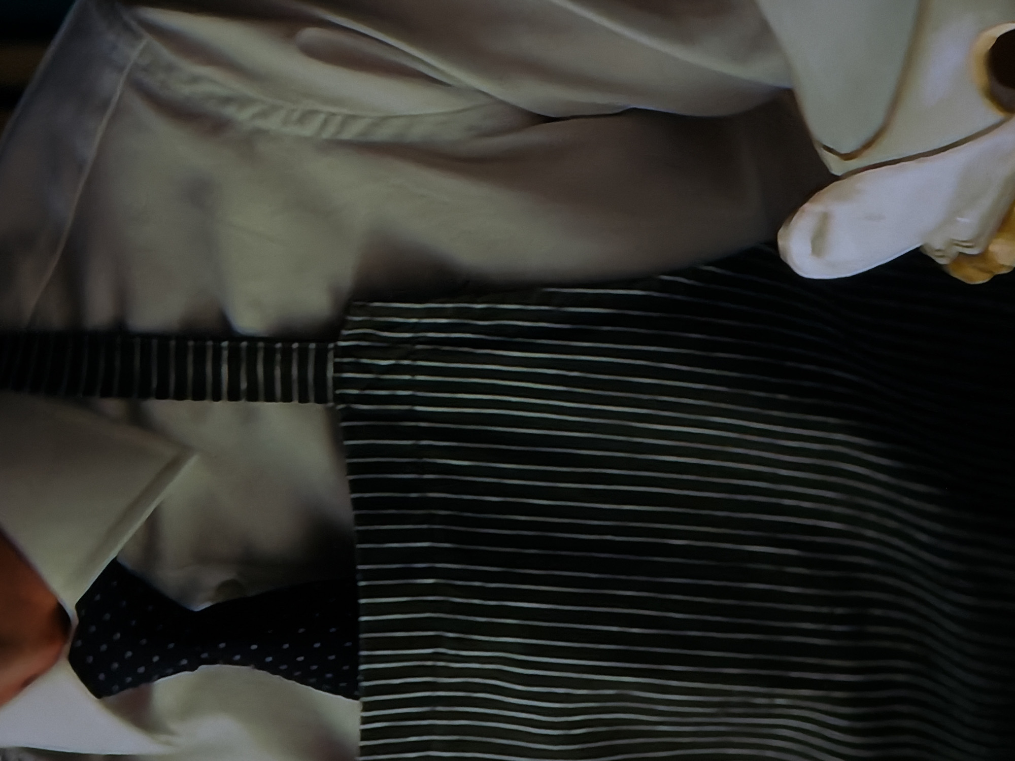 A close-up, high-angle shot captures a person's upper torso and clothing, with an unknown object partially visible on the right. The person is wearing a light, possibly cream or beige, long-sleeved dress shirt. A dark bow tie with small white polka dots is visible at the collar, neatly tied. Over the shirt, a dark garment with thin, distinct white horizontal stripes covers the lower part of the frame, resembling an apron or a piece of striped fabric. This striped garment extends across the person's front.
To the upper right of the frame, a segment of a white or cream-colored object with gold-colored trim is visible. It appears to be a stylized form, possibly part of a doll's or mannequin's head or hand, with rounded edges and a small opening or detail. The lighting is soft, creating gentle shadows on the shirt fabric and accentuating its folds. No specific activity is depicted, and the scene appears to be static. The image is a detailed view of textiles and a sculptural element, with no visible text or direct environmental cues to the location or time of day.