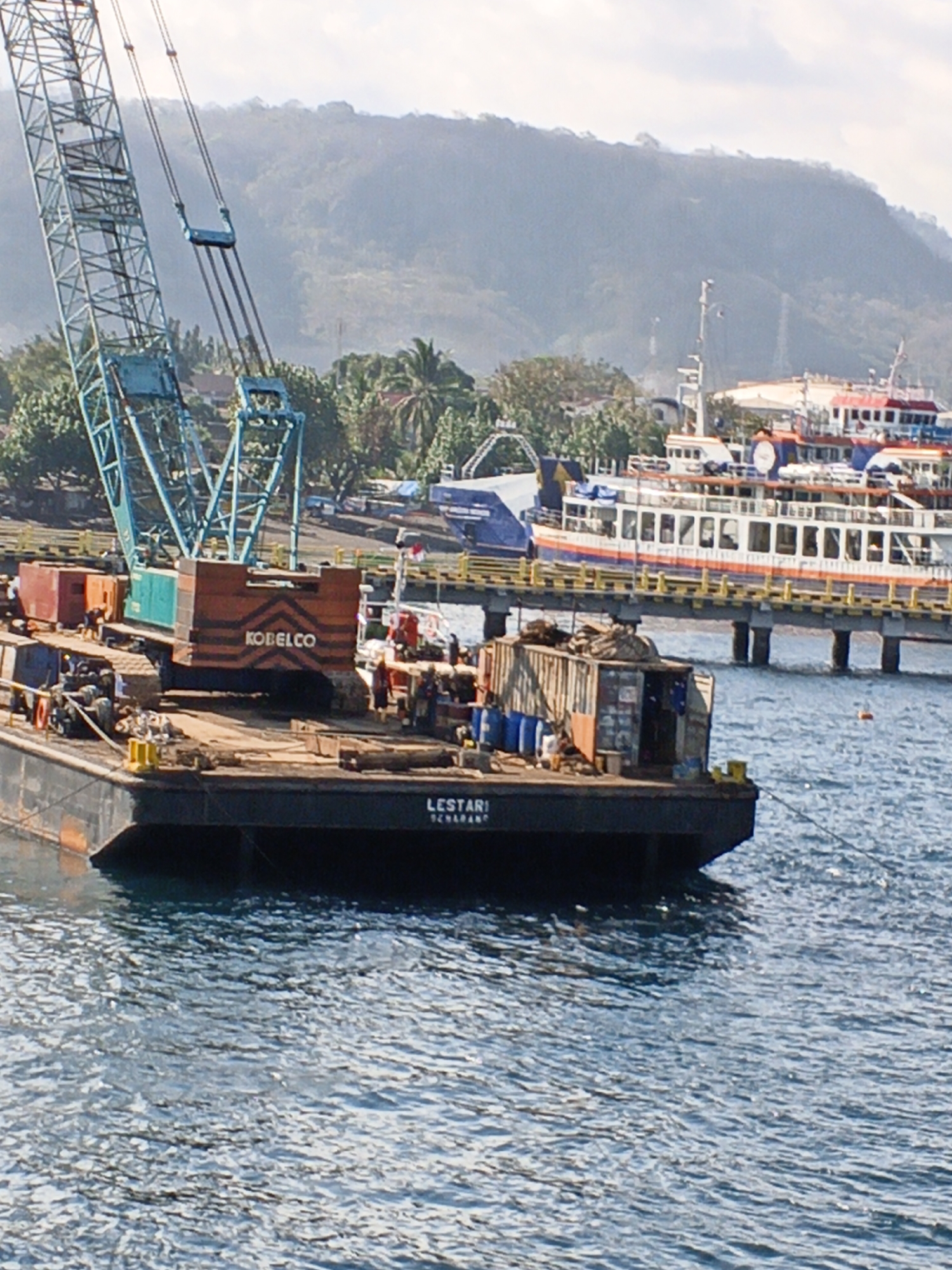 The image shows a large barge floating in the water near a pier in Banyuwangi, Indonesia. On the barge, there is a large blue crane and several shipping containers. The crane has the word "KOBELCO" written on its side. The barge also has "LESTARI SCHAARNDAM" written on its side. In the background, there are several large ferries docked at the pier and a lush, green hillside with trees and buildings. The sky is cloudy.