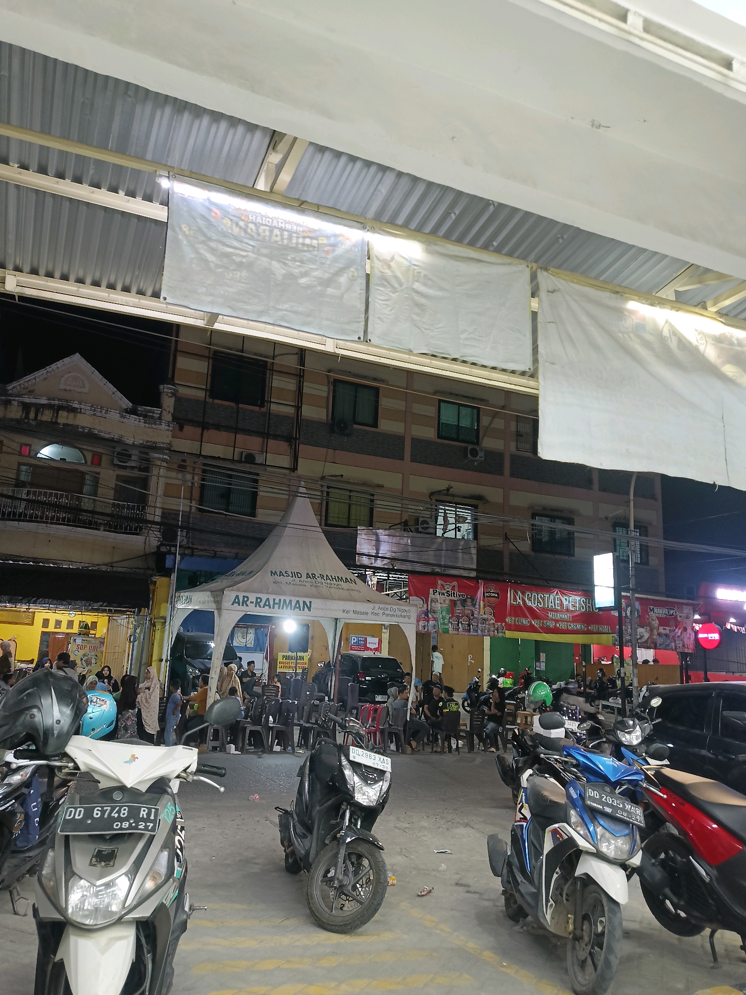 This is a medium-distance, eye-level shot of a street scene at night in Makassar, Indonesia, likely in the Panakkukang area given the presence of multiple businesses with Indonesian signage. In the foreground, three scooters are parked. The leftmost scooter is a light-colored automatic model with the license plate "DD 6748 RI". The middle scooter is a darker automatic model, partially obscured. The rightmost scooter is a blue and white automatic model with the license plate "DD 2035 KAR".
Behind the scooters, a white tent with the prominent text "AR-RAHMAN" and "MASJID AR-RAHMAN" is set up. Underneath the tent, people are seated at tables, suggesting a gathering or dining area. To the left of the tent, a building with multiple floors is visible, with some windows illuminated. Further to the left, a yellow building with a white gable is seen.
On the right side of the frame, more businesses with various signs are displayed, including "LA COStatistical PETSHOP" and "MARKOTOPS." Red lights and neon signs indicate ongoing commercial activity. The overall environment appears to be a bustling street with both residential and commercial elements, lit by artificial lights. The presence of parked vehicles and pedestrian activity suggests a common gathering spot for locals.