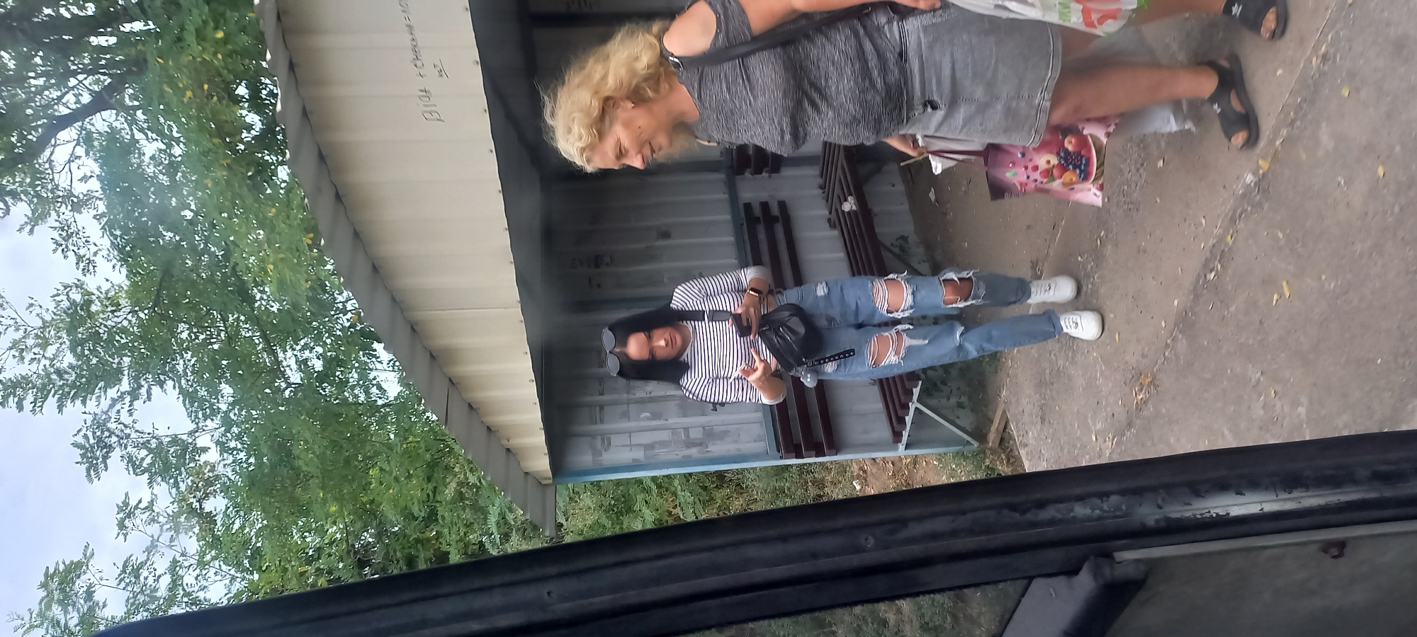 Two women are standing at a bus stop. The woman on the left is young, with long dark hair, wearing a striped long-sleeved shirt and ripped jeans. She is holding a smartphone and has a fanny pack around her waist. The woman on the right is older, with blonde, curly hair, wearing a grey t-shirt and a grey denim skirt. She is carrying shopping bags. The bus stop has a light-colored roof and benches. The area is surrounded by trees and vegetation. The bus stop is located in Klyntsi, Ukraine.