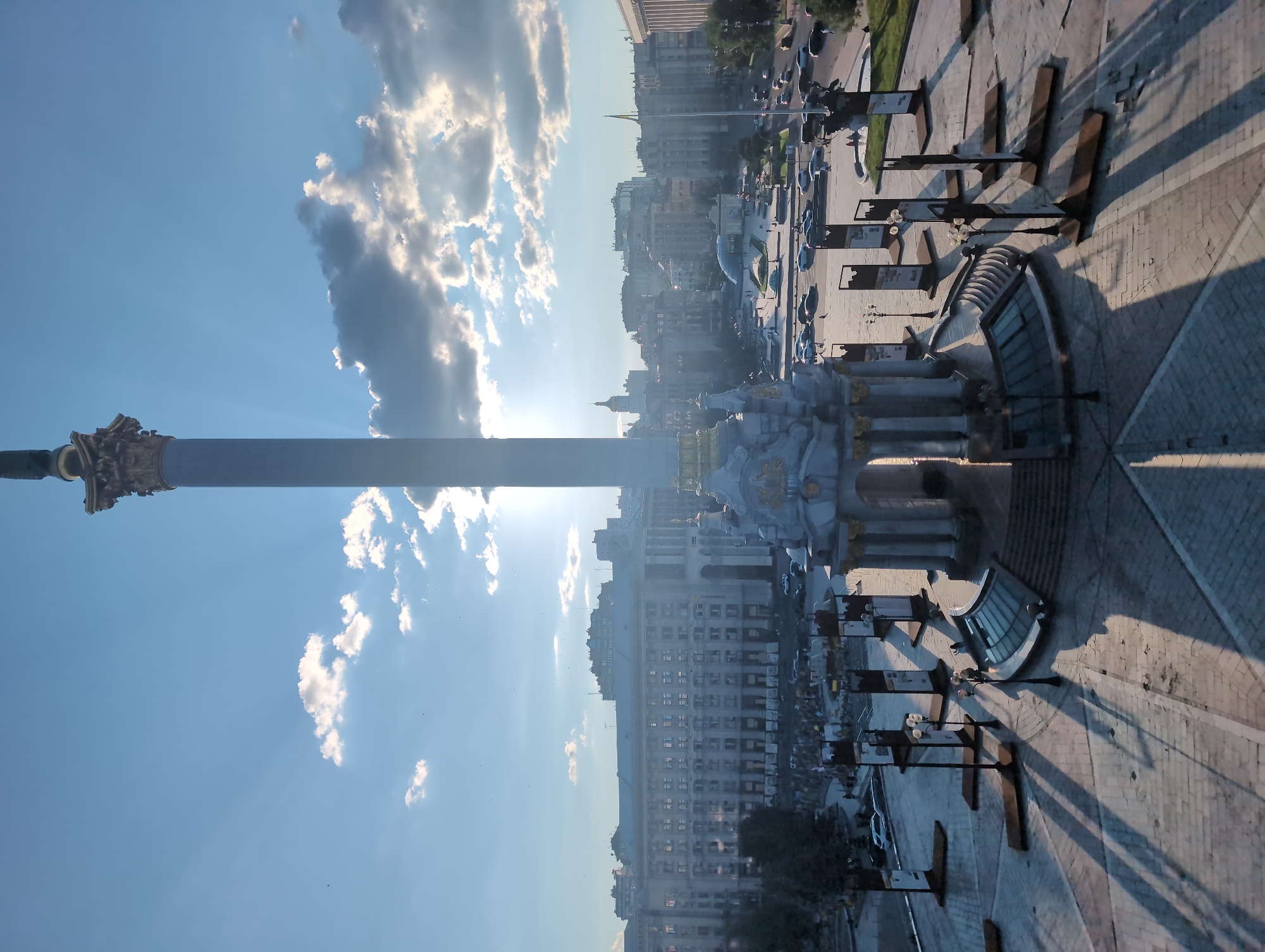 This is a wide-angle, high-angle shot of Maidan Nezalezhnosti (Independence Square) in Kyiv, Ukraine, taken during daylight with a clear blue sky and some scattered clouds. The sun is positioned behind a tall column monument, casting long shadows across the paved square. The monument itself is ornate, with a large column topped by a statue. Surrounding the monument is a circular base with architectural detailing. In the foreground, there are several large, rectangular display stands with dark frames.
In the midground, there is a street with moderate traffic, including several cars and what appear to be buses. A distinctive curved glass structure is visible, and further back, a long, grand building with many windows dominates the skyline. Several other buildings of varying heights and architectural styles are visible in the distance, forming the city's urban landscape. A Ukrainian flag is visible on a flagpole to the right of the frame. People are present in the scene, some walking in the square and others visible near the buildings and in the street. The overall atmosphere suggests a public space with ongoing activity under a bright sky.