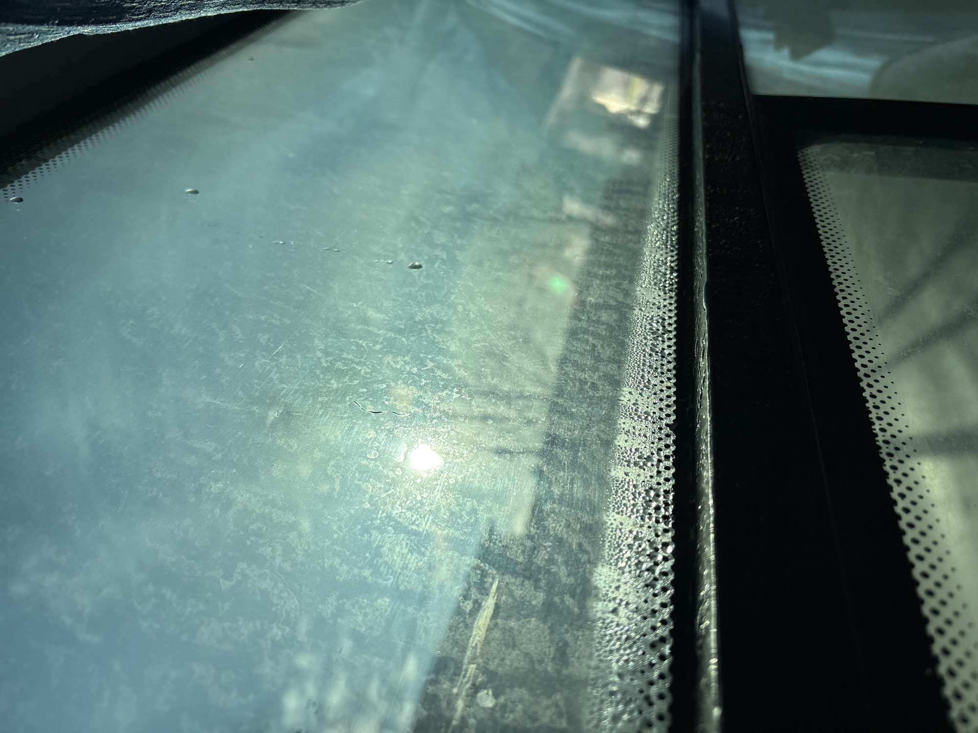 The image shows the interior view through a dirty, possibly steamed-up window, with condensation visible along the edges. A dark-colored frame divides the view into sections. The sun is visible through the window, creating a bright glare. The location in Berezniki, Russia, cannot be determined from the image alone.