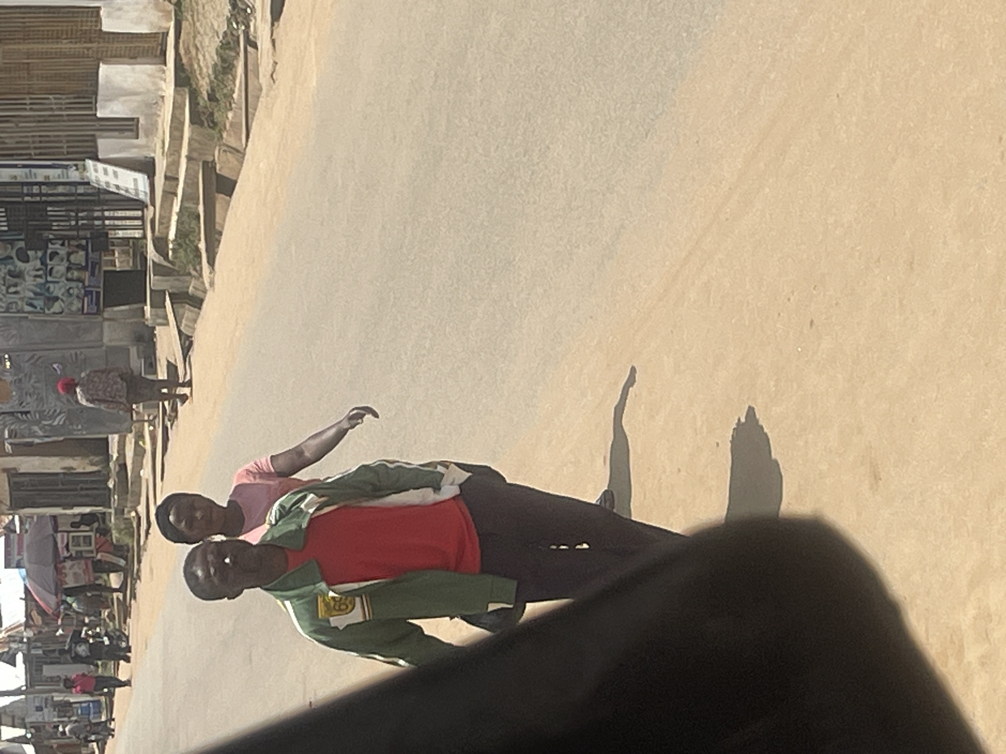 The image depicts a street scene in Abuja, Nigeria. In the foreground, a person wearing a green jacket over a red shirt and dark pants walks towards the viewer, with a partial view of the person from the chest down and their left side. Behind them, a woman in a pink shirt and dark pants also walks in the same direction. The street is paved with asphalt and appears to be lined with buildings and stalls on the left side. Some people are visible further down the street, and there is a market stall with an umbrella. The environment appears to be an urban or semi-urban setting.