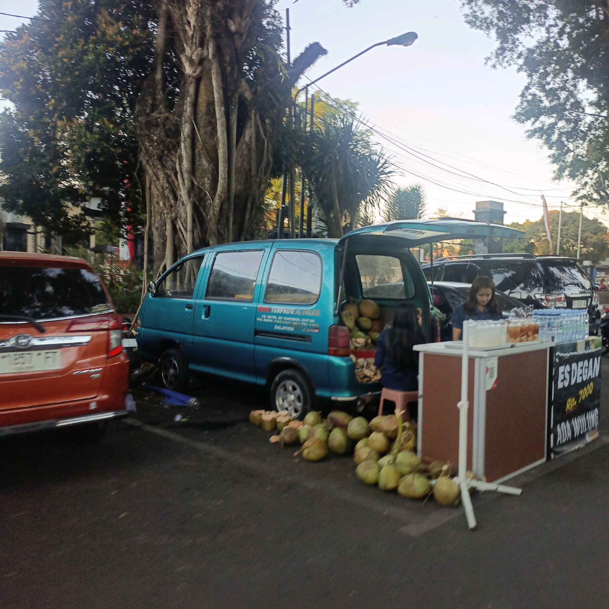 This image depicts a street scene in Salatiga, Indonesia, with a focus on a beverage stand and parked vehicles. In the foreground, a red SUV is parked on the right side of the frame. To its left, a teal-colored van is parked, with its rear cargo door open, revealing a large quantity of coconuts. A woman is standing near the back of the van, and another person, possibly a customer, is seated on a small stool.
In the background, a street vendor's stall is set up, featuring drinks, bottled water, and a sign that reads "ES DEGAN" and "Rp. 7000". Behind the stall, a black SUV is parked. The environment includes large trees with exposed roots, a street lamp, and power lines, suggesting an outdoor urban setting. The ground is paved, and there are a few scattered coconuts on the road. The lighting indicates it is likely daytime, possibly late afternoon or early morning due to the warm tones.