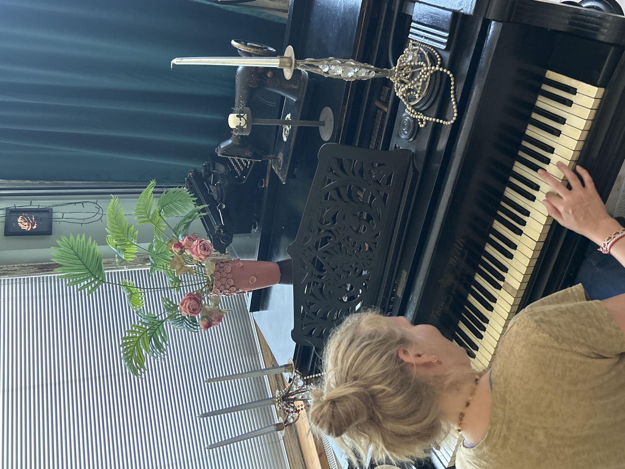 In an indoor scene, a blonde-haired woman with her hair in a bun is sitting in front of a black piano, playing it. She is wearing a tan t-shirt and jeans, and a necklace. On the piano, there is a book stand, a vintage sewing machine, a typewriter, a vase with artificial pink roses and palm leaves, two candelabras, and a string of pearls. The background features a window with horizontal blinds and a dark green curtain. The floor is not visible.