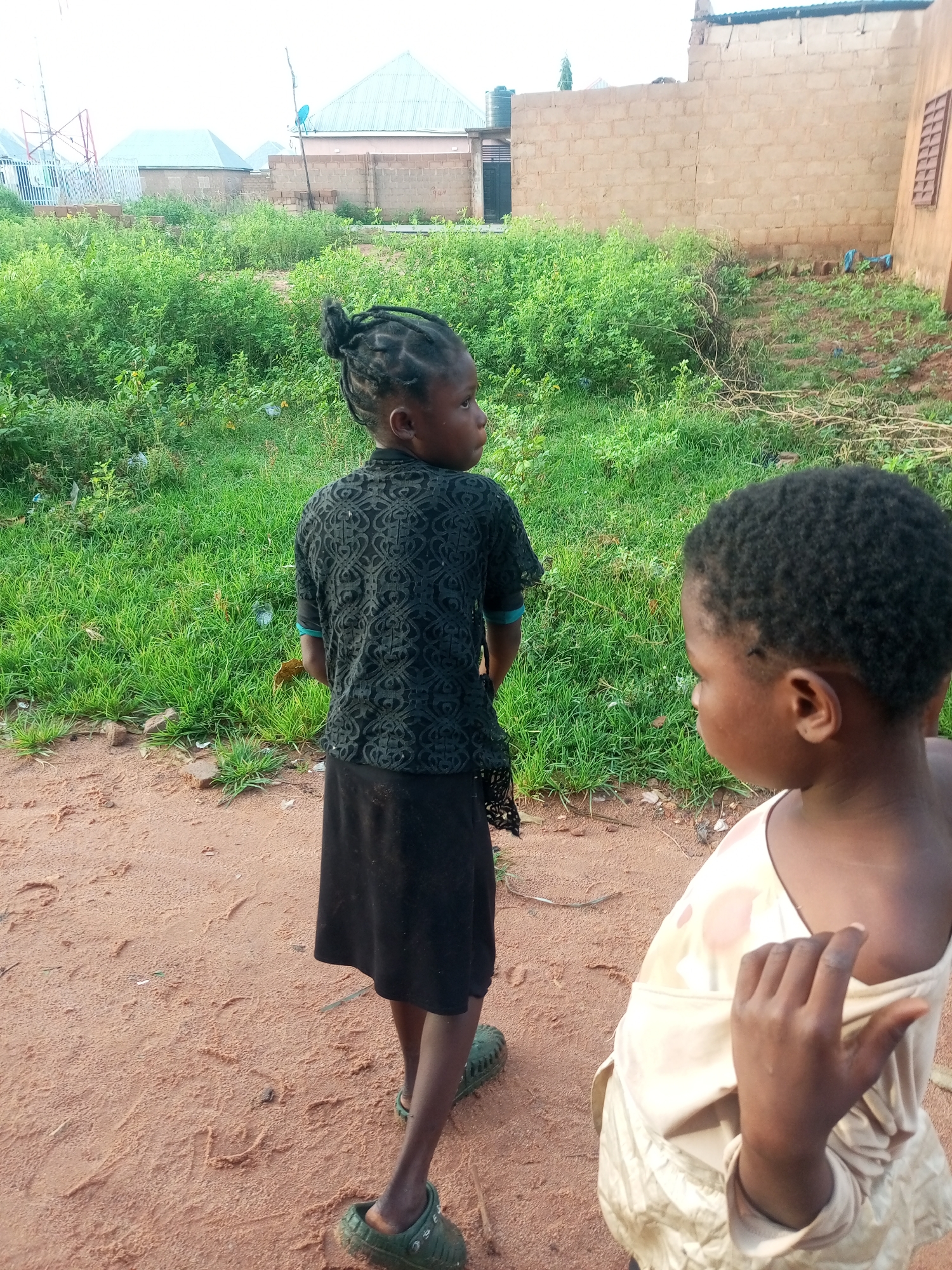 The image depicts two young girls standing on a dirt path in what appears to be a semi-urban or rural setting in Tempure, Nigeria. The girl in the foreground on the left is facing away from the camera, with her back towards the viewer. She has dark skin, her hair is styled in braids pulled into a bun, and she is wearing a black, short-sleeved top with an intricate pattern and a black skirt. She is also wearing green clogs.
To her right and slightly in front of her is another young girl, also with dark skin. She is facing towards the left of the frame, with her profile visible. Her hair is short and dark. She is wearing a light-colored garment, possibly a dress or top, with a patterned design in shades of pink and cream. Her right hand is raised and resting on the fabric of her clothing.
The environment is characterized by a dirt path with some scattered debris and footprints. Behind the girls, there is an area of dense green vegetation, likely overgrown grass and bushes. In the background, several buildings are visible. On the left, there are structures with light-colored roofs, one appearing to be a house with a light blue satellite dish on top, and another with a pale green water tank. Further in the background, a tall, metal structure, possibly a communication tower, stands out. To the right, a building constructed with beige-colored bricks is prominent, with a small, dark-paned window visible on its side. The sky is overcast, suggesting a cloudy or hazy day. The overall impression is a scene of everyday life in a developing community.