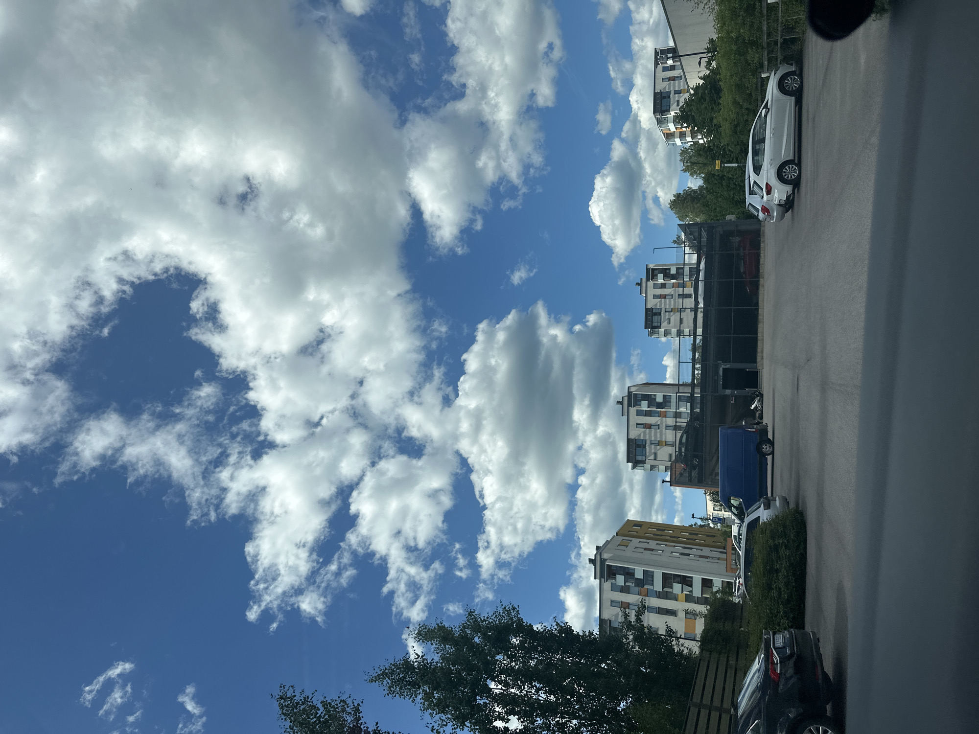 A photograph captures a daytime scene in an urban or suburban environment. The sky is a vibrant blue, dotted with scattered white cumulus clouds. In the foreground, a dark-colored car is partially visible, likely from the driver's perspective. Beyond the car, a paved parking lot is present, with several other vehicles parked, including a silver SUV, a blue van, and a white hatchback. A multi-story parking structure with dark-colored railings stands in the middle ground. Behind the parking structure, modern, multi-story apartment buildings with colorful facades are visible. Lush green trees and bushes are interspersed throughout the scene, suggesting a well-maintained outdoor space. The overall impression is one of a contemporary residential or commercial area on a clear day.