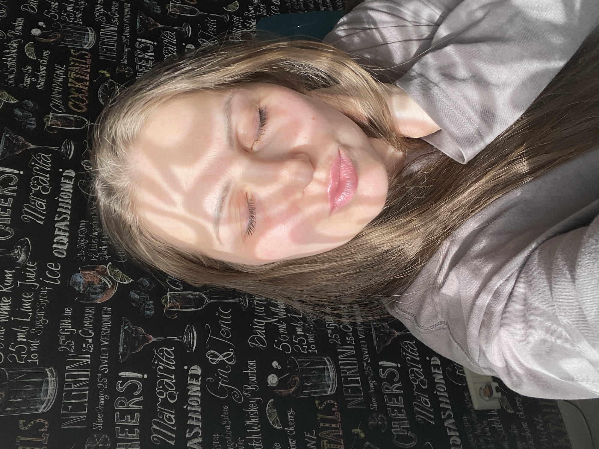 A young woman is depicted resting or sleeping, with her eyes closed, illuminated by strong, dappled sunlight. The light casts distinct shadow patterns across her face, suggesting she is positioned near a window on a sunny day. She has long, light-brown hair spread around her and is wearing a plain grey long-sleeved top. Her lips have a light pink hue, possibly from lip gloss or lipstick.
She is lying on a dark surface, which appears to be a tablecloth or a tabletop, intricately patterned with white illustrations and text. This pattern features various cocktail recipes, ingredients, and measurements, giving the impression of a bar or pub-themed design.
Visible text on the surface includes the names of cocktails such as "NEGRONI," "Margarita," "Gin & Tonic," "Old Fashioned," "Champagne Cocktails," and "Daiquiri." Specific ingredients and measurements like "25ml Gin," "25ml Campari," "Slice Orange," "Sweet Vermouth," "60ml Scotch Whiskey Bourbon," "Lime Juice," and "Sugar syrup" are also clearly readable. The celebratory word "CHEERS!" is repeated across the pattern. The overall ambiance is peaceful and relaxed, enhanced by the natural light. This scene is set in Markovskii, Russia.