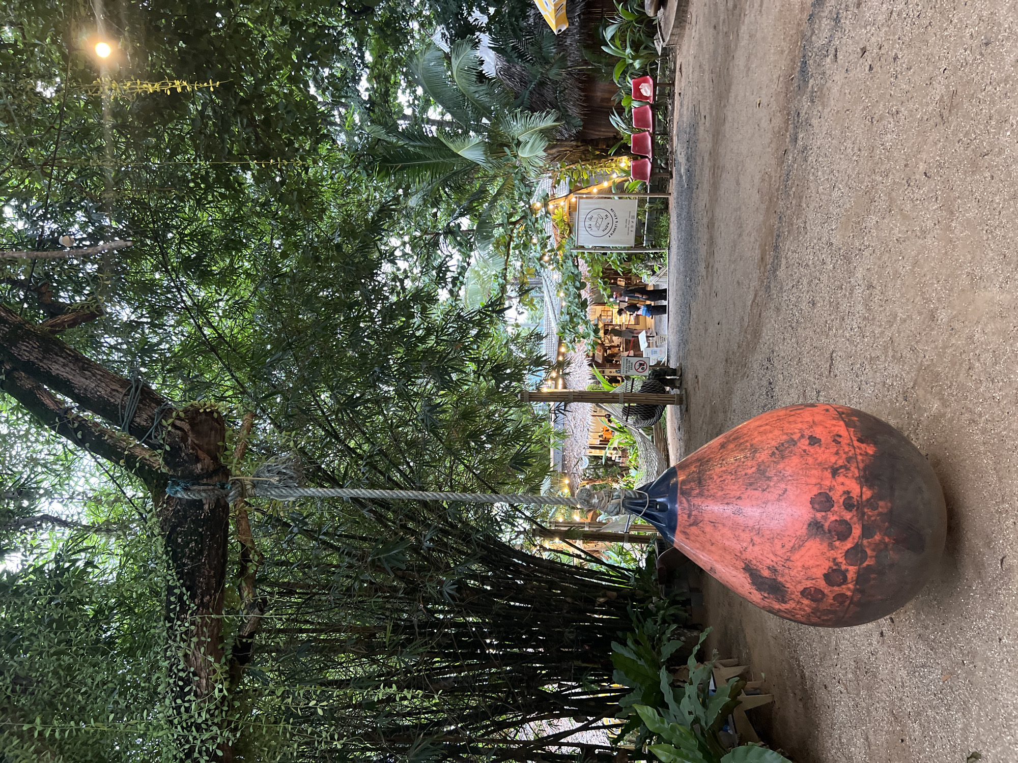 This image captures an outdoor scene at what appears to be a cafe or restaurant in Karon, Thailand. In the foreground, a large, orange, tear-drop shaped object, likely a buoy, hangs from a rope attached to a tree branch, partially obscuring a grove of bamboo on the left. The ground is a mix of dirt and gravel. In the mid-ground, a pathway leads towards a cluster of buildings with thatched roofs, illuminated by string lights. Several red chairs are lined up outside one of these structures. A white sign with Thai writing and the English text "A BLANKET & A PILLOW" is prominently displayed. A few people are visible near the buildings, and a hammock can be seen hanging between two wooden posts. Lush green foliage and trees surround the area, creating a tropical ambiance. The lighting suggests it is late afternoon or early evening.