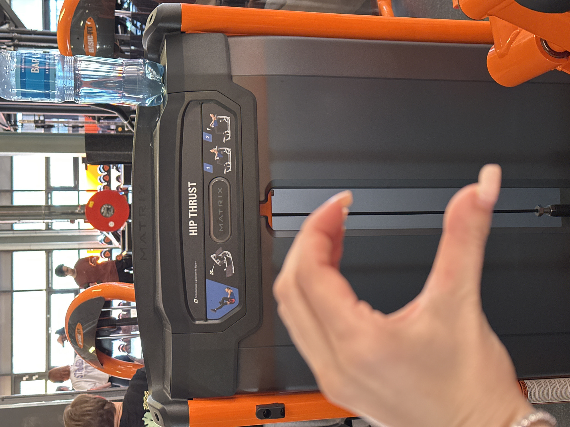 This media file captures a dynamic indoor scene at a gym, likely in Amsterdam, Netherlands. The central focus is a "MATRIX HIP THRUST" exercise machine, which is predominantly dark gray with bright orange structural elements.
In the midground, a plastic water bottle with a blue label for "BAR LE DUC NATUUR WATER" rests on the machine's console. Adjacent to the bottle, the machine's control panel features instructions for use, including two diagrams labeled "1" and "2" depicting the proper body position for the hip thrust exercise. Below these diagrams, there's text that reads "Selectionnez la position de départ" (French for "Select the starting position"). The "MATRIX" brand name is visible in two places on the machine.
The foreground shows a person's light-skinned hand, with manicured nails and a ring on one finger, slightly out of focus on the lower right, suggesting the photo was taken by someone actively using or observing the gym.
The background reveals more of the gym environment. Several individuals are present, some appearing to be exercising or moving between machines. One person with dark hair is visible standing in a brown shirt, and another with lighter hair is partially seen wearing a white shirt. Other gym equipment, including an orange piece with "BASIC-FIT" branding and a red circular weight plate, is discernible. Large windows in the background let in natural light, suggesting it is daytime.
The presence of "BAR LE DUC" water (a Dutch brand) and "BASIC-FIT" (a gym chain prominent in the Netherlands and other European countries), coupled with the provided location context of Amsterdam, firmly places the scene in a fitness facility in the Netherlands.