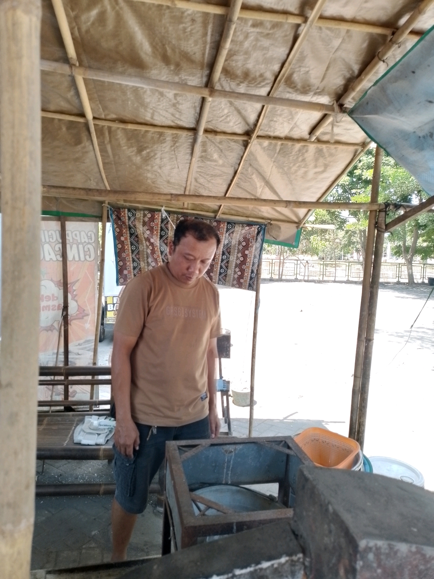A man is standing in what appears to be an open-air food stall or temporary shelter. The shelter has a roof made of bamboo poles covered with a translucent material. The man is wearing a light brown t-shirt with some text on it and dark shorts. He is looking down. In the foreground, there is a metal structure, possibly a cooking stove or display. To the man's left, there is a bench made of bamboo with some white items on it. Behind the man, there are two banners or signs, one with red and white text and a graphic. Further in the background, there are trees, a fence, and a bright, sunny outdoor area. The location is identified as the city of Wonosari, Indonesia.