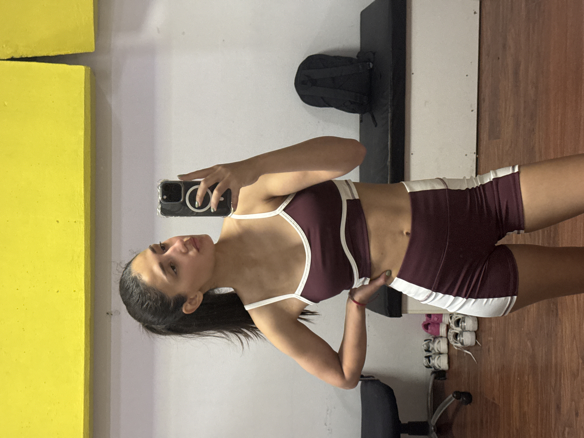 A woman is taking a selfie in what appears to be a gym or fitness studio. She is wearing a maroon and white sports bra and matching shorts. Her hair is pulled back in a ponytail. The background includes a yellow structure, a white wall, a black bench, a backpack, a collection of shoes, and a chair. The location, Bar, Montenegro, is not evident from the image.