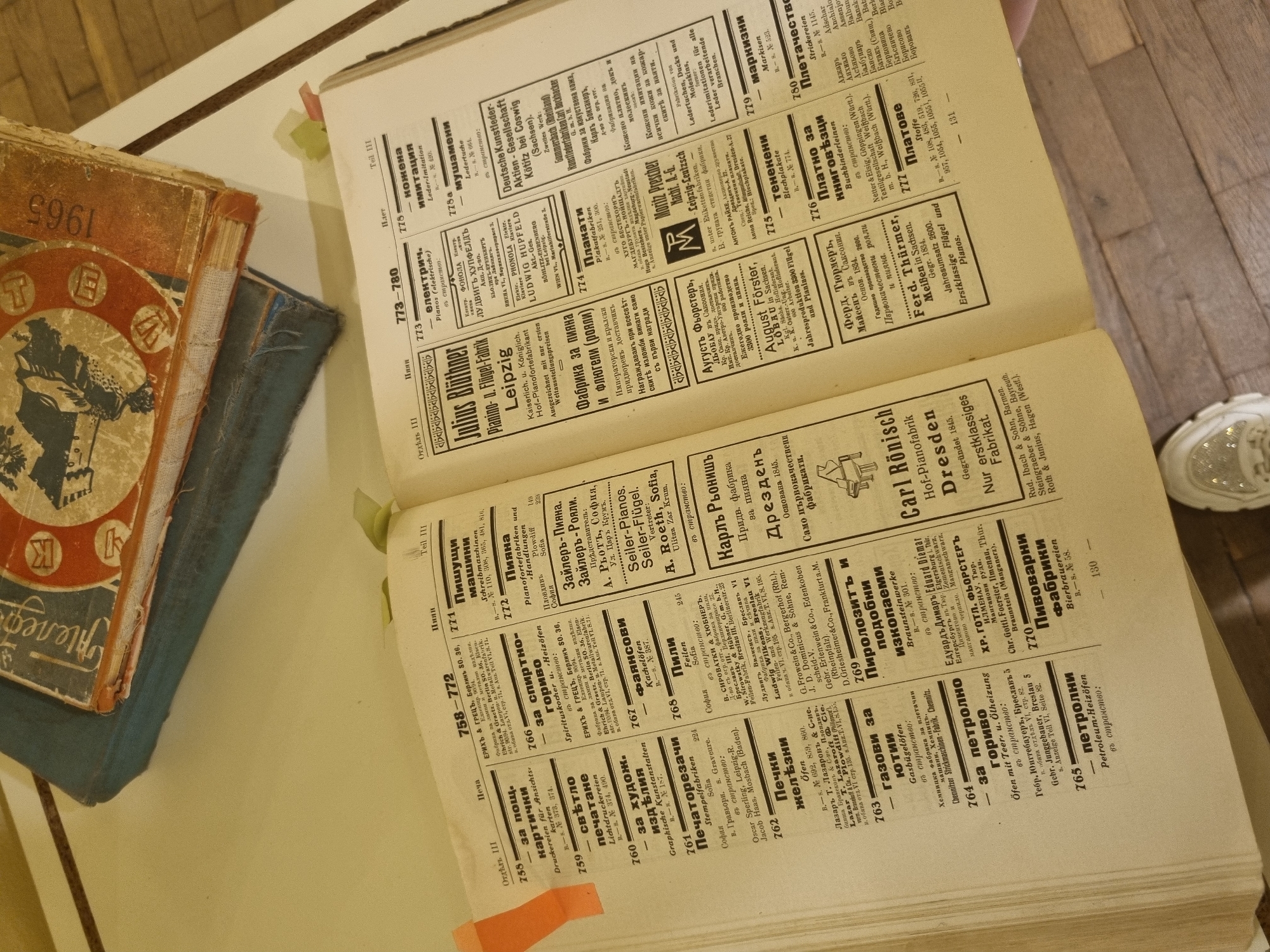 The image shows an open book with a yellowed page and faded print. The pages are filled with text in a Cyrillic-based script, likely Bulgarian or a related language. On the left side of the image, there's a stack of old, worn books with faded covers. The topmost book has a distinct design featuring a figure and the year 1965. The book contains information for the city of Gorna Oryahovitsa, Bulgaria.