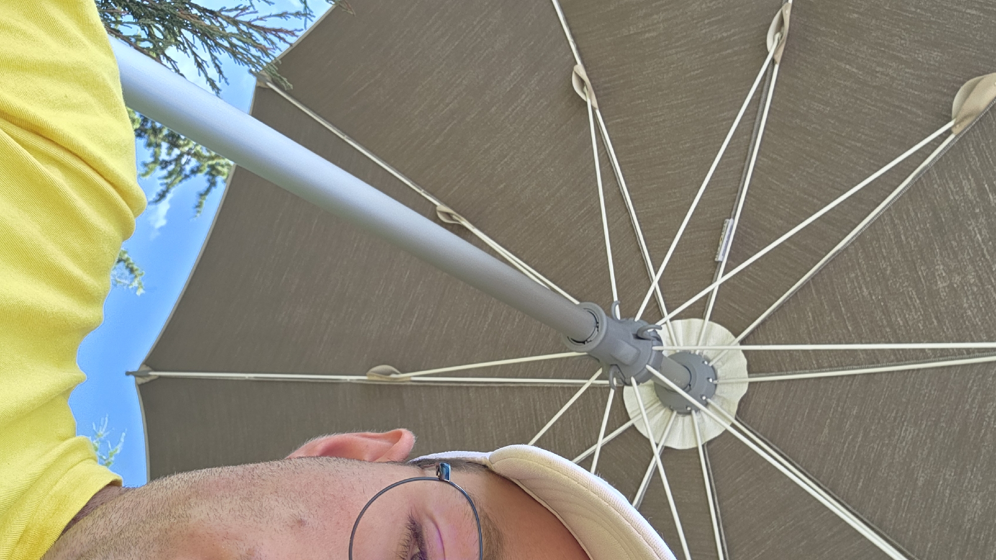 A close-up, low-angle shot shows the underside of a large, brown umbrella, its spokes radiating outward. A person's head and shoulder are visible on the right side of the frame, wearing a light-colored baseball cap and round-rimmed glasses. The person's right eye is visible. A bright yellow shirt is worn on the shoulder. The sky is blue with some clouds visible, and green foliage can be seen in the background, suggesting an outdoor setting, likely in a park or garden. The lighting suggests a bright, sunny day.