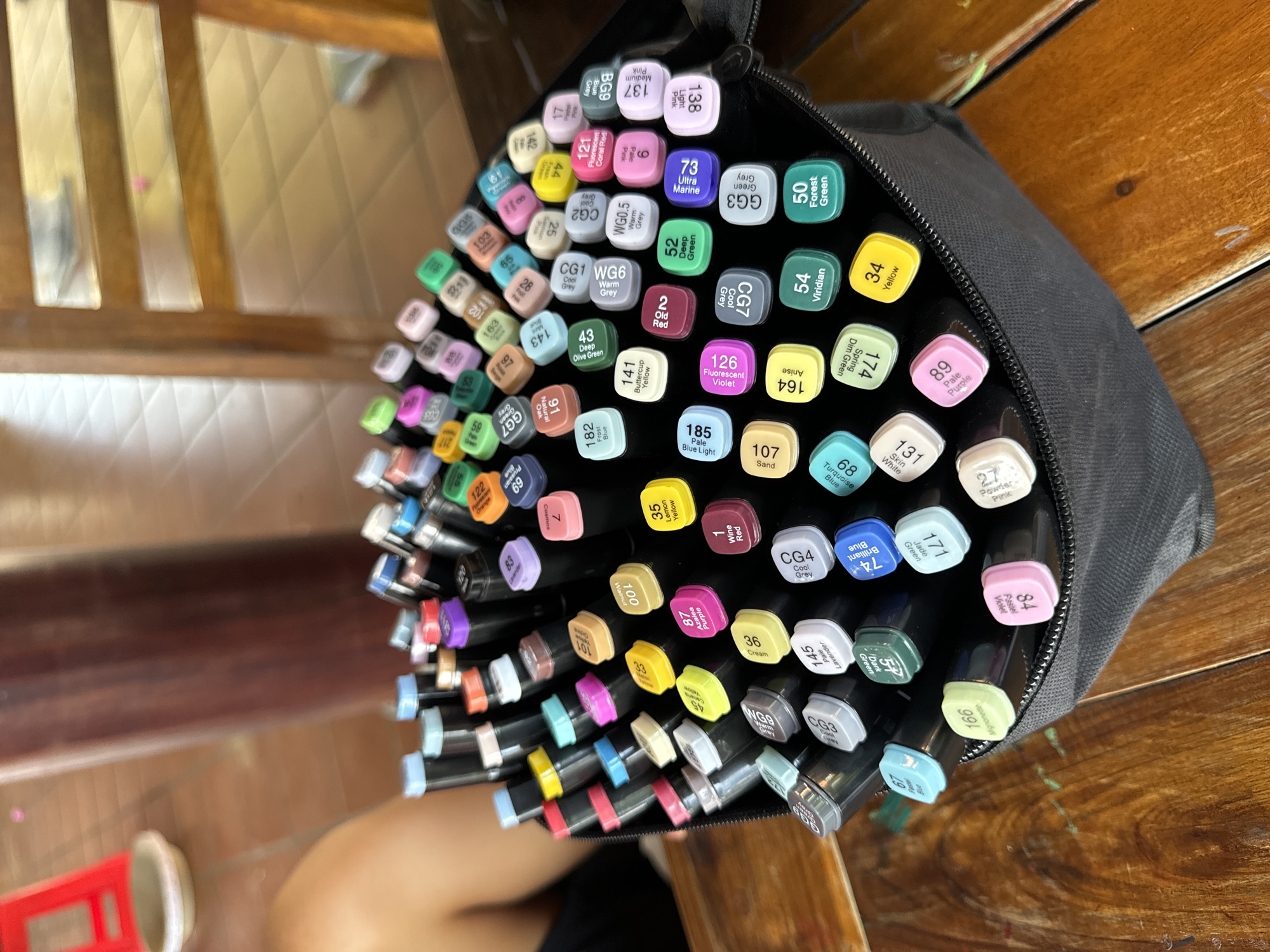 A top-down shot depicts a black zippered pouch filled with numerous colorful markers, likely alcohol-based, arranged haphazardly. Each marker has a cap displaying a color name and number. The pouch sits on a wooden surface. A chair with a metal frame and red plastic seat is visible in the lower left corner. The scene is indoors, with a glimpse of another chair in the background. The location is presumed to be in Mũi Né, Vietnam, based on context, but there are no elements in the photo to confirm that. No people are clearly visible in the frame.