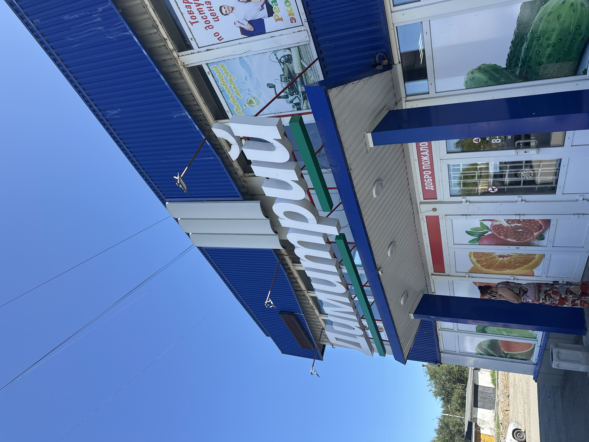 The image shows the exterior of a blue building with large white 3D letters spelling out "Дмитрий" (Dmitry) above the entrance. The building appears to be a store, with large windows displaying images of fruits and vegetables like watermelon, oranges, and pomegranates. A woman in a patterned dress is standing outside the entrance, talking on her phone. A silver car is parked to the left of the building. The sky is clear and blue, and power lines are visible against it. The building is situated in an urban or semi-urban area, with a concrete structure and trees visible in the background. The presence of Cyrillic text indicates the location is likely in a Russian-speaking country. Based on the provided information, this scene is in Biysk, Russia.