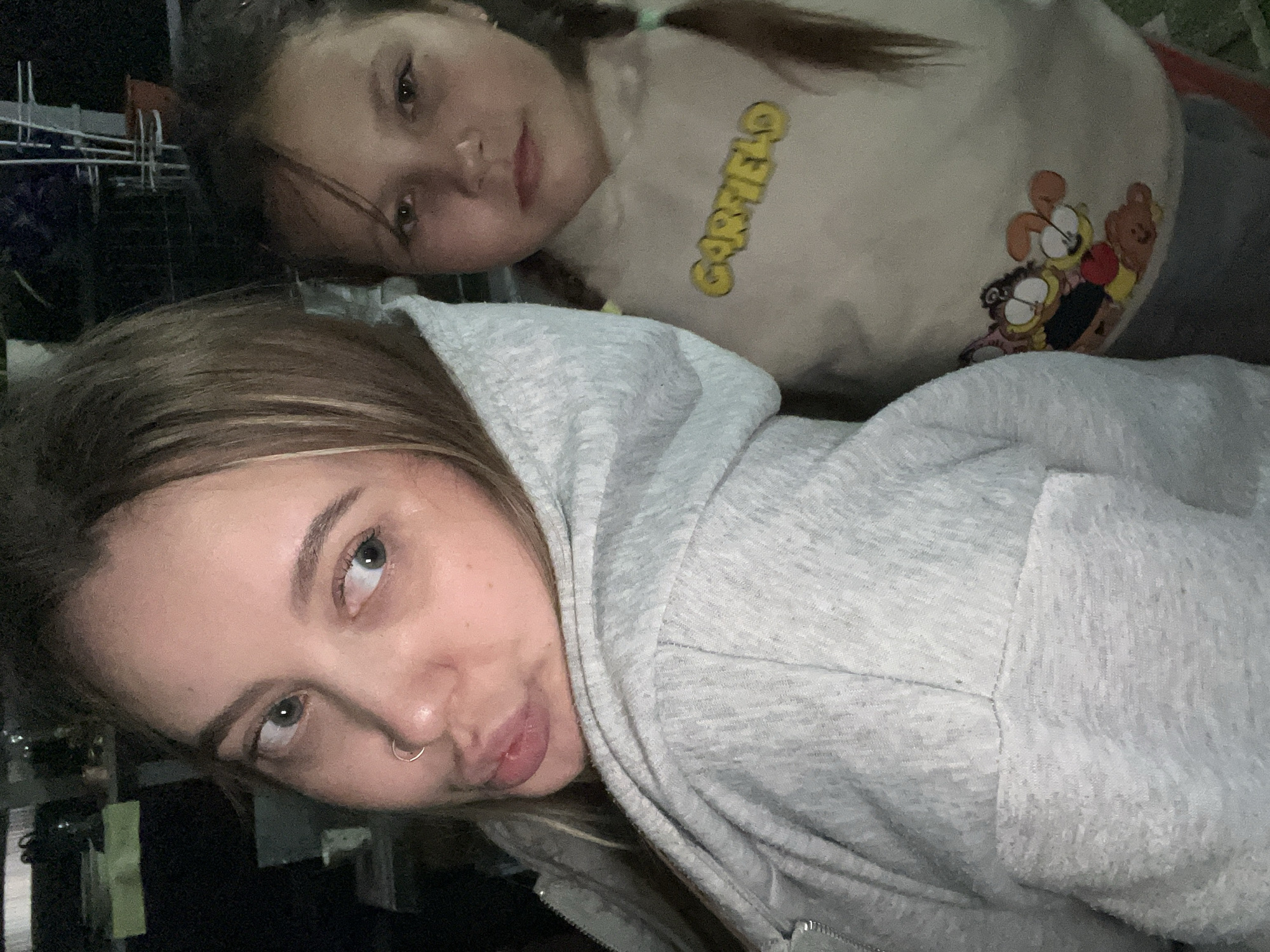 Two individuals are captured in an indoor selfie. The foreground features a woman with light brown hair, a nose ring, and wearing a light grey hooded sweatshirt. Behind her, slightly out of focus, is a young girl with braided hair and wearing a beige sweatshirt with a cartoon graphic depicting the word "GARFIELD" and associated characters. The location is identified as Novopokrovskaya, Russia. The lighting is subdued.