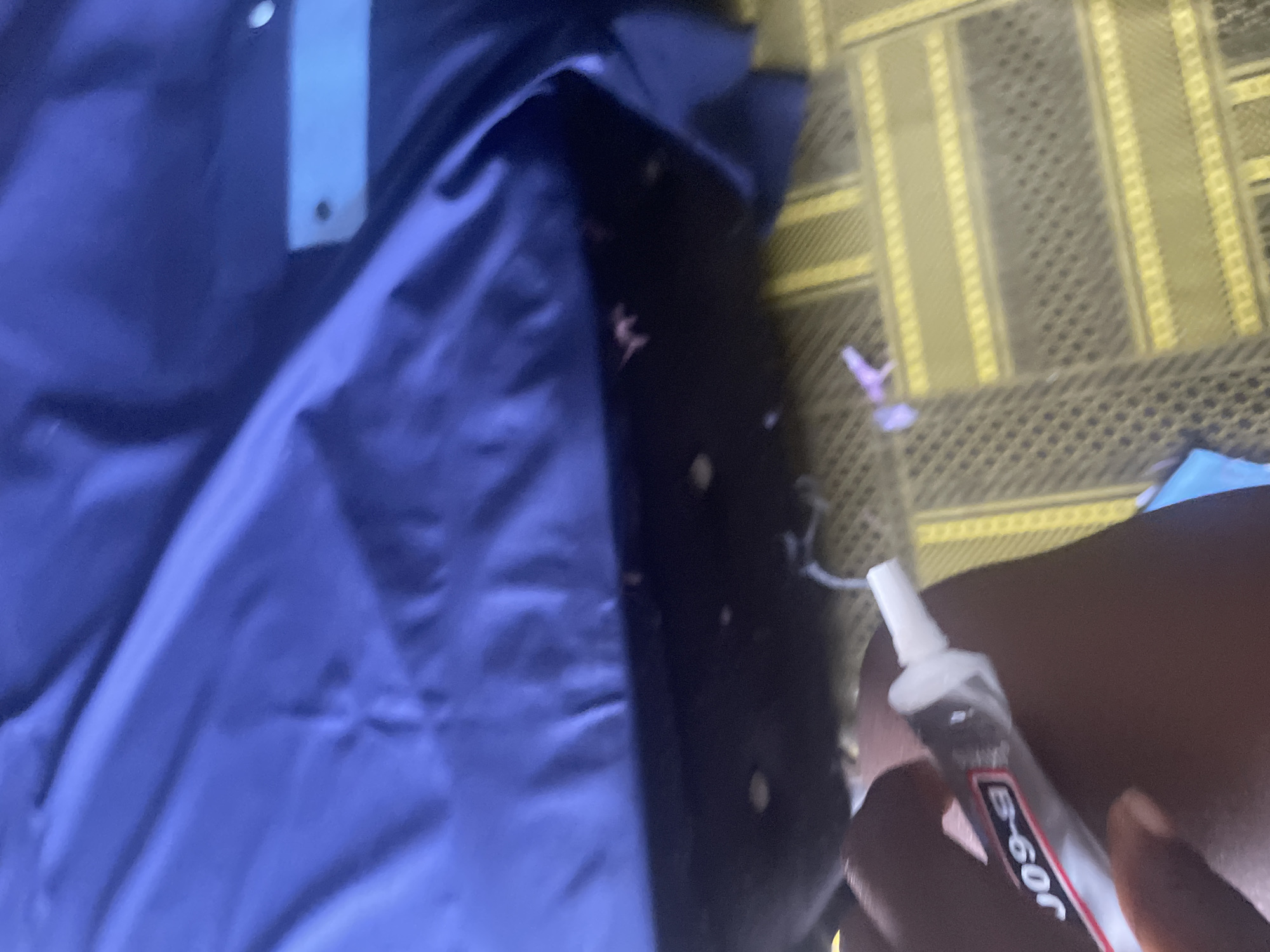 In the city of Jalingo, Nigeria, a close-up shot captures a person's hand holding a tube of glue. The tube is white with red and black lettering that reads "B-60R". The glue tube is partially transparent, revealing some of the adhesive inside. The person's dark skin is visible against the backdrop of a large, dark blue, possibly synthetic fabric, which appears to be a piece of clothing or upholstery. The fabric has visible folds and texture. Below the blue fabric, a patterned surface is visible, characterized by yellow and dark brown stripes and a grid-like texture, suggestive of a mat or carpet. A small, light purple object, possibly a piece of plastic or fabric, is also visible on this patterned surface. The overall lighting suggests an indoor setting, possibly a room.