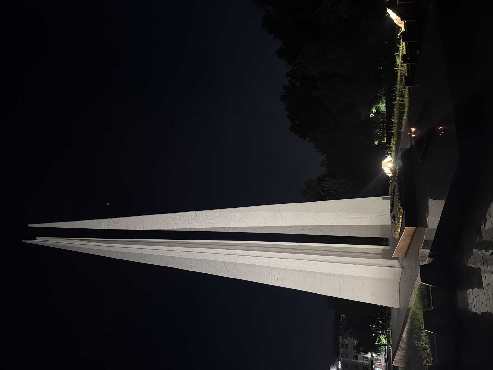 This is a nighttime photograph of the Eternal Flame monument in Pavlodar, Kazakhstan. The monument consists of three tall, white, obelisk-like structures that taper to a point, positioned close together. The central and left obelisks appear to be slightly taller and wider than the right one. At the base of the monument, there is a circular or oval relief sculpture, possibly depicting a scene or a symbol. Around the base, there are what appear to be eternal flames or gas torches burning, casting a warm glow. The monument is situated on a raised platform with a reflective surface in the foreground, which appears to be a water feature or a polished stone that reflects the monument and the flames. Beyond the monument and to the right, there is a line of dark, silhouetted trees. To the left of the monument, there is a building with illuminated windows and a sign that is partially visible. The sky is dark, indicating nighttime. The overall scene is solemn and commemorative.