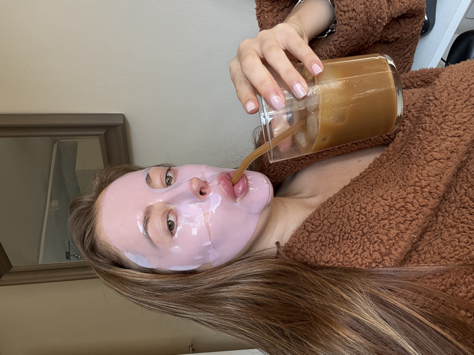 A close-up, indoor photograph taken in the city of Unknown, Unknown, shows an individual, identified as female, from the chest up. She is wearing a translucent pink sheet face mask that covers her forehead, cheeks, nose, and chin, leaving her eyes and mouth exposed. Her light brown hair is visible framing her face. She is dressed in a textured, fuzzy brown garment, likely a robe or loungewear. The individual is actively drinking from a clear glass filled with iced coffee, using a light brown, curved straw. Her right hand, with nails manicured and coated in a clear or light pink polish, holds the glass. In the background, a plain light-colored wall features a rectangular mirror with a dark wooden frame, reflecting a ceiling light fixture and a light-toned interior surface. A small, light-colored fixture is partially visible on the far left.