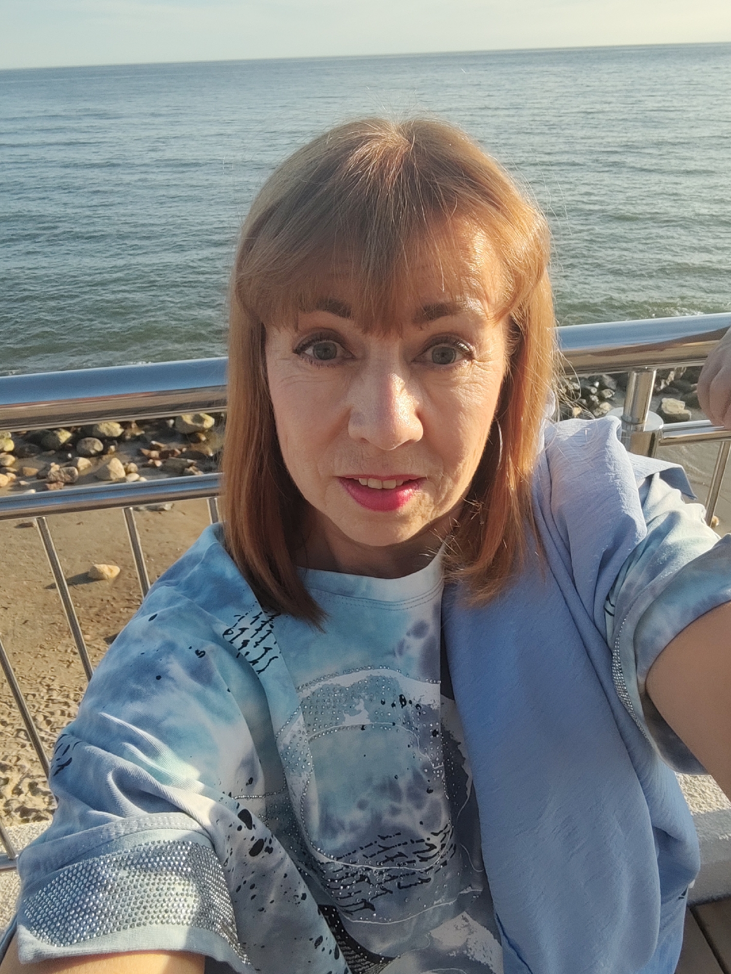 An outdoor, eye-level, waist-up, selfie image shows a light-skinned Caucasian woman with shoulder-length auburn hair and bangs standing outdoors with the ocean and sky in the background. She is looking directly at the camera with her mouth slightly open and her eyes wide. She has bright pink lipstick on and is wearing a light blue tie-dye shirt with black and white patterns, some of which are adorned with rhinestones. She also has a light blue scarf draped over her shoulders. She is standing behind a metal railing that separates her from a sandy beach with large rocks. The scene appears to be in Svetlogorsk, Russia, as indicated by the context of previous images, suggesting a coastal location.