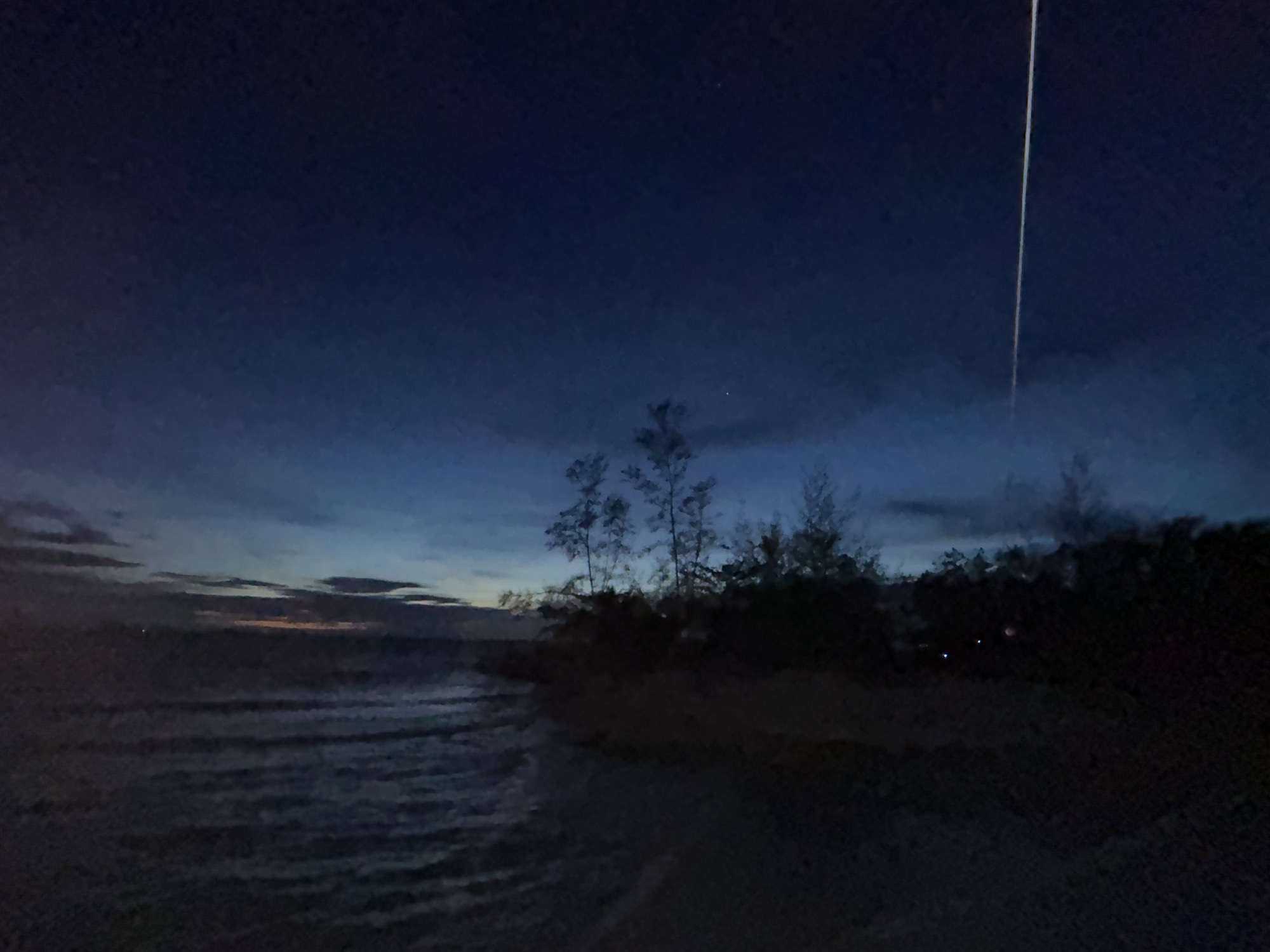 The image depicts a coastline at twilight or night. To the left, waves can be seen approaching a dark shoreline. Further inland, a line of trees and shrubs are silhouetted against the sky. A streak of light is visible vertically in the upper right portion of the sky. The location is Baan Sri Thanu, Thailand.