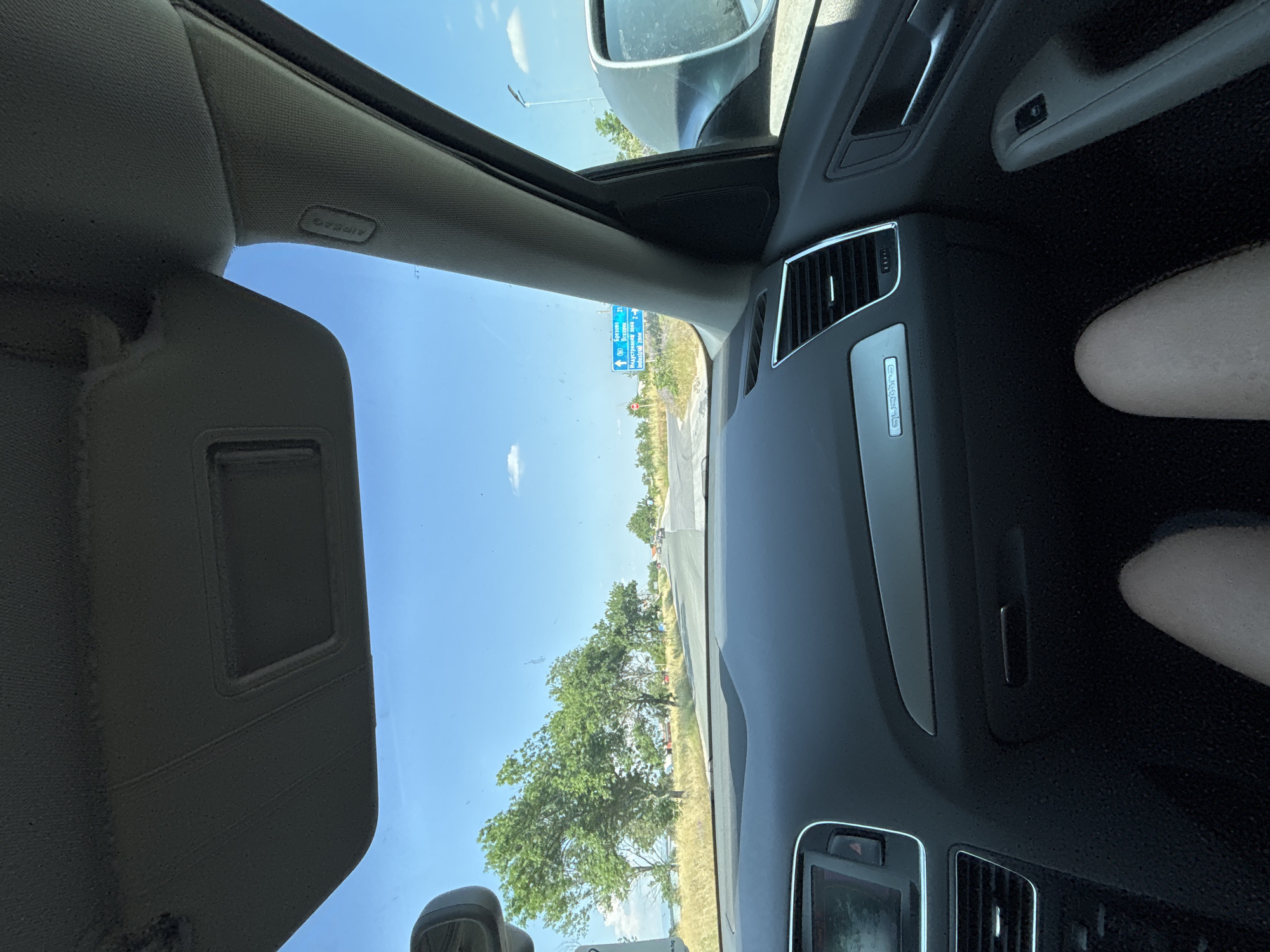 An interior shot from a car, rotated 90 degrees counter-clockwise, shows the view from the passenger's seat looking towards the front and passenger side. The car appears to be an Audi, evidenced by the "Quattro" badge on the dashboard. Visible interior elements include a beige sun visor, the headliner with an "AIRBAG" label on the A-pillar, air vents, a section of the door panel, and the passenger's legs.
Through the clear windshield and passenger window, a bright, clear blue sky with a few wispy white clouds is visible. The external scene features a road or highway with green trees and dry grassy areas along the side. In the distance, a blue road sign with white text is clearly legible, indicating directions to "Бургас" (Burgas) and "КАРНОБАТ" (Karnobat), along with "21" and "2006" (likely distances in kilometers), and "Индустриална Зона" (Industrial Zone). A red circular sign with the number "6" inside is also visible at the top of the blue sign, suggesting a road number.
The image captures a sunny, daytime scene, likely during travel or a stop, in the vicinity of Stryama, Bulgaria, given the Bulgarian text on the road sign pointing towards Burgas and Karnobat. The weather is bright and clear.