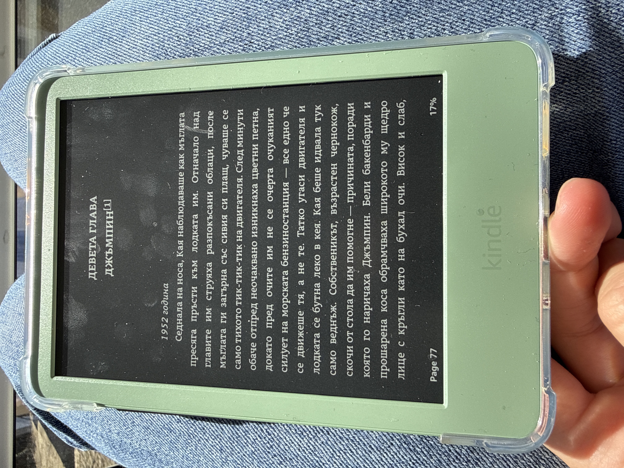 Here is a description of the image:
The image shows a Kindle e-reader displaying a page of text in Bulgarian. The device is encased in a clear green protective case and rests on what appears to be a denim-clad surface, possibly a lap. The text displayed is an excerpt from a novel, with the title "ДЕВЕТА ГЛАВА" (Ninth Chapter) and a name "ДЖЪМПИН" (Jumping). The chapter begins with the year "1952 година" (1952 year) and describes a scene where the character Kaya observes the fog as their boat approaches a marine gas station. The text continues to describe the station and its owner, an elderly black man with white sideburns and a kind face. At the bottom of the screen, "Page 77" and the Kindle logo are visible, along with a battery indicator showing 17%.