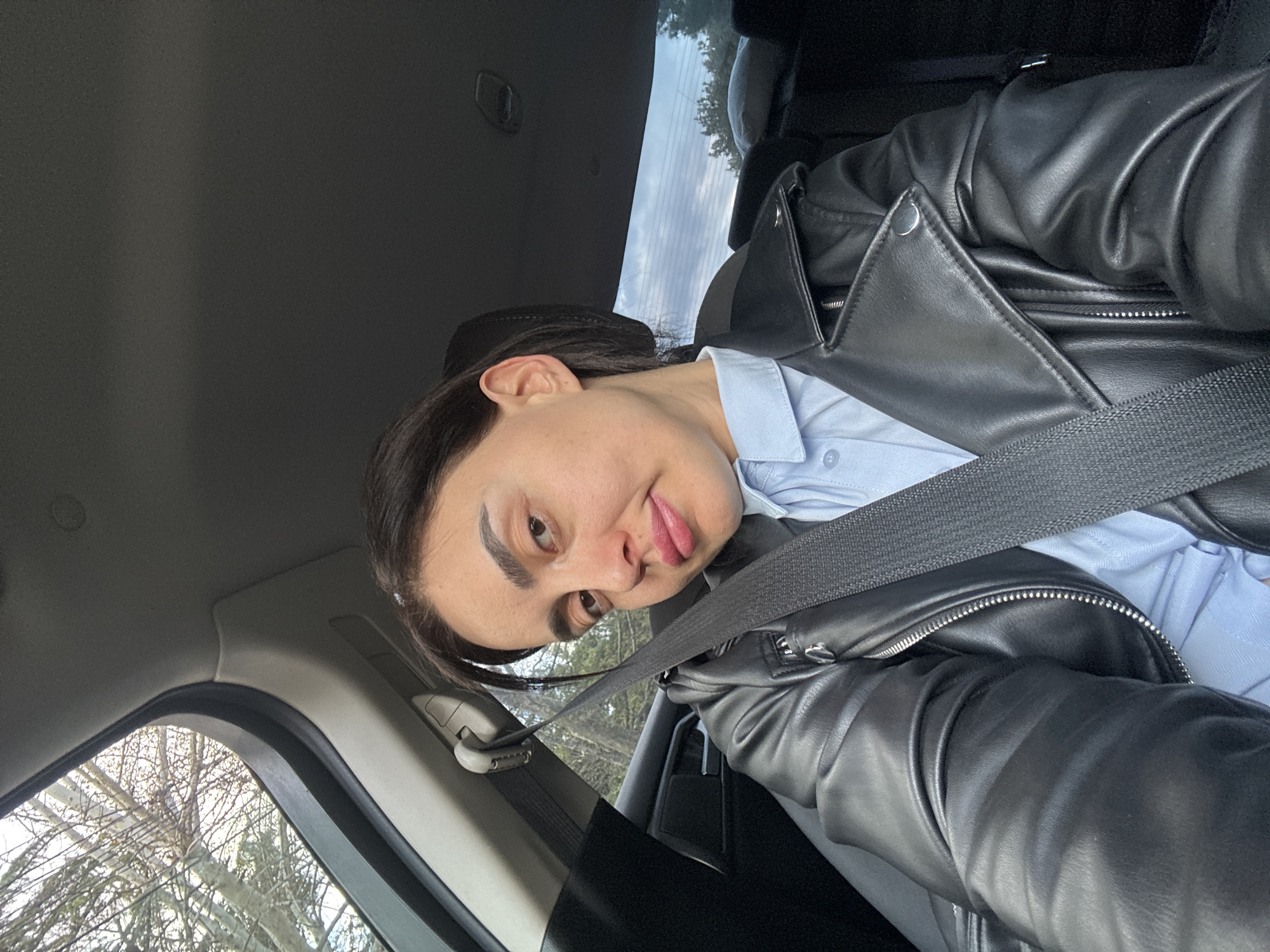 A woman is seated in the driver's seat of a vehicle, wearing a light blue collared shirt and a black leather jacket. A seatbelt is secured across her chest. She is looking at the camera with her tongue slightly sticking out. The interior of the car is visible, with part of the window frame and some trees visible outside the window. The car's ceiling is a light gray color. The city and location are not determinable from this picture.