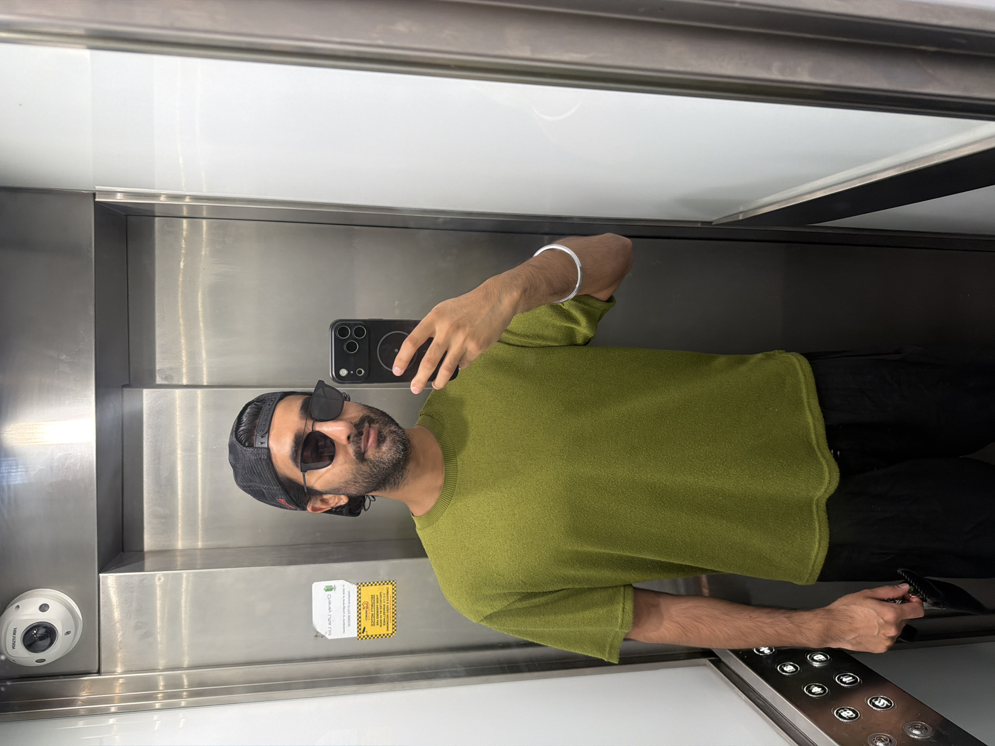 A man takes a selfie in an elevator. He is wearing a green t-shirt, black shorts, and a black baseball cap. He is also wearing sunglasses and a silver bracelet on his left wrist. He is holding his phone in his right hand. The elevator appears to be in a modern building, possibly a hotel or office building. There is a security camera mounted on the ceiling of the elevator. The elevator walls are made of stainless steel, and there are buttons on the left wall. A yellow sticker with text in English is visible on the left wall of the elevator. The man is of South Asian descent, with dark hair and a beard. He is smiling faintly at the camera. The overall setting is clean and well-lit.