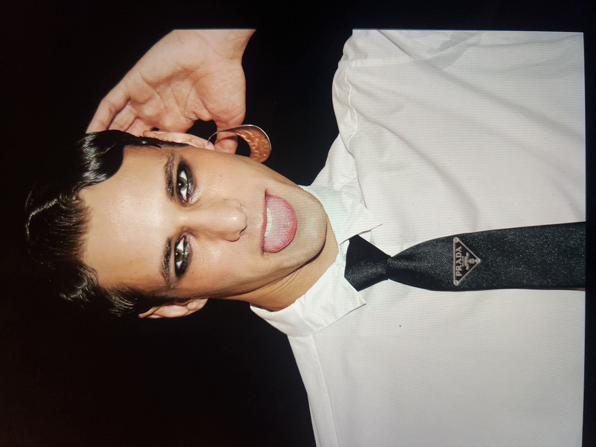 The image shows a close-up, head-and-shoulders shot of a person with dark, slicked-back hair styled with finger waves. They are wearing a white collared shirt and a black tie with a triangular Prada logo. The person has heavy eye makeup and is sticking out their tongue, with their right hand touching their ear. They are wearing a large, crescent-shaped earring. The background is dark and out of focus. The image appears to be taken indoors. There is no information to indicate this is in a specific city or country.